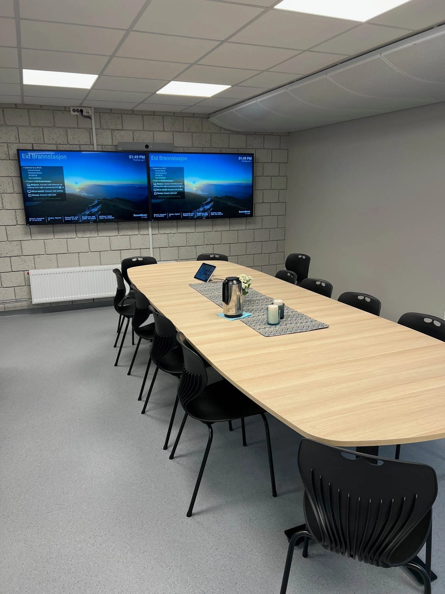 Et konferanserom med en lang bord med stoler rundt, to digitale skjermer på veggen som viser naturbilder, og et bord med kaffekanne, kopper og notatblokker.