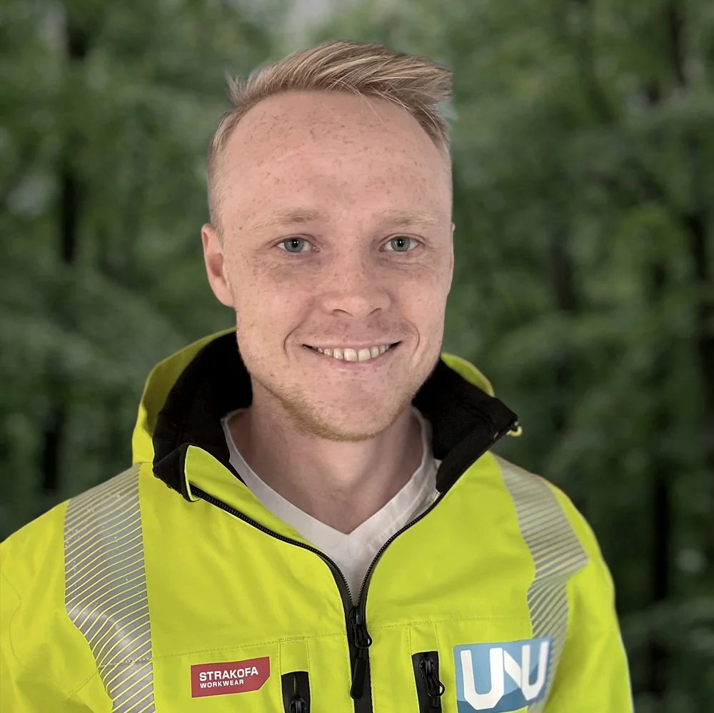 En ung mann med lyst hår og fregner smiler, iført en gul refleksvest med logoer, stående foran en grønn skog.
