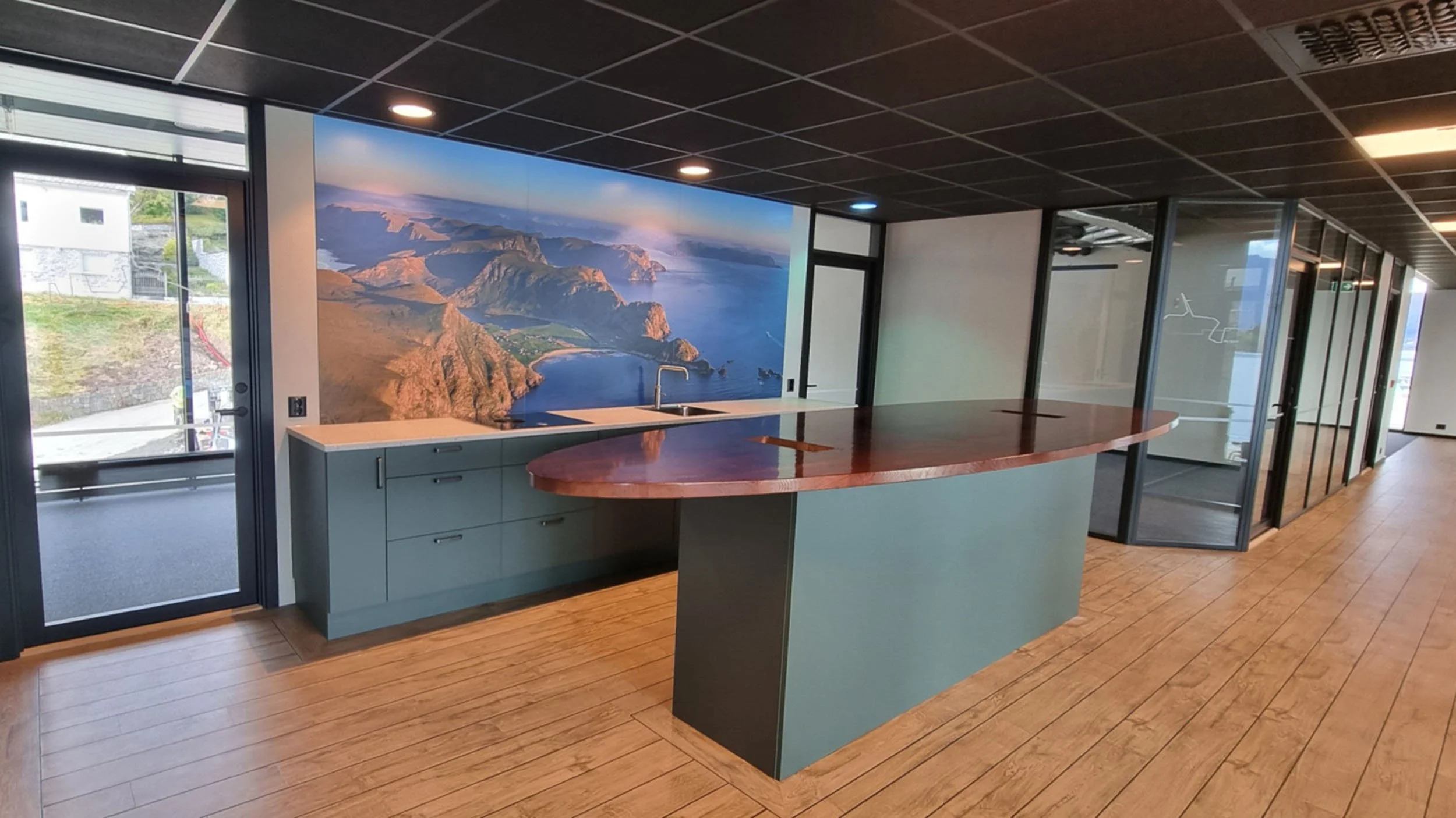 Interiør av et kontor med en moderne kjøkkenøy, store vinduer og et fjell- og sjølandskap dekket av en veggdekorasjon
