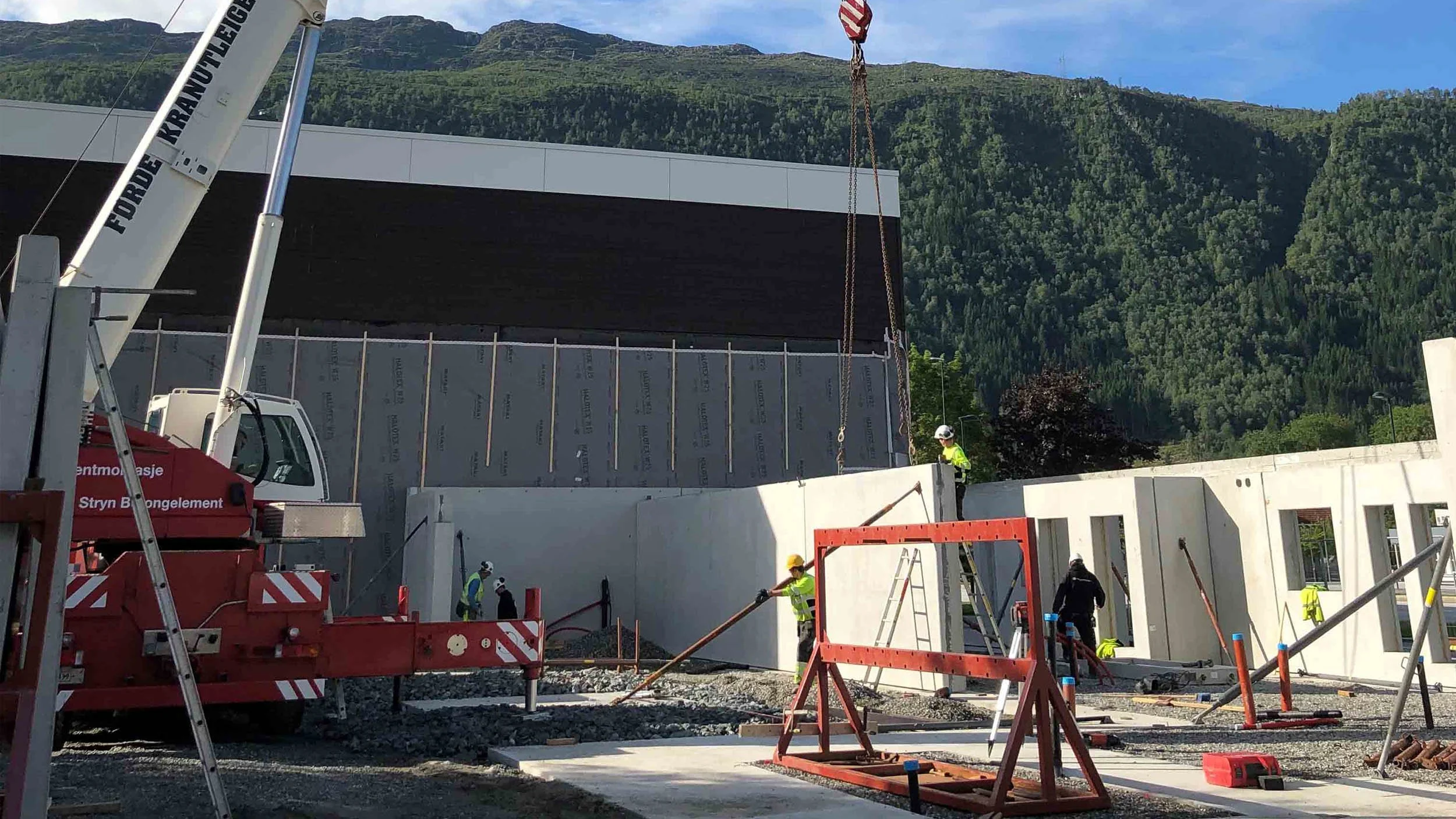 Byggearbeidere i høyt synkronisert arbeid på en byggeplass med fjell i bakgrunnen, og en kran som løfter et stort element.