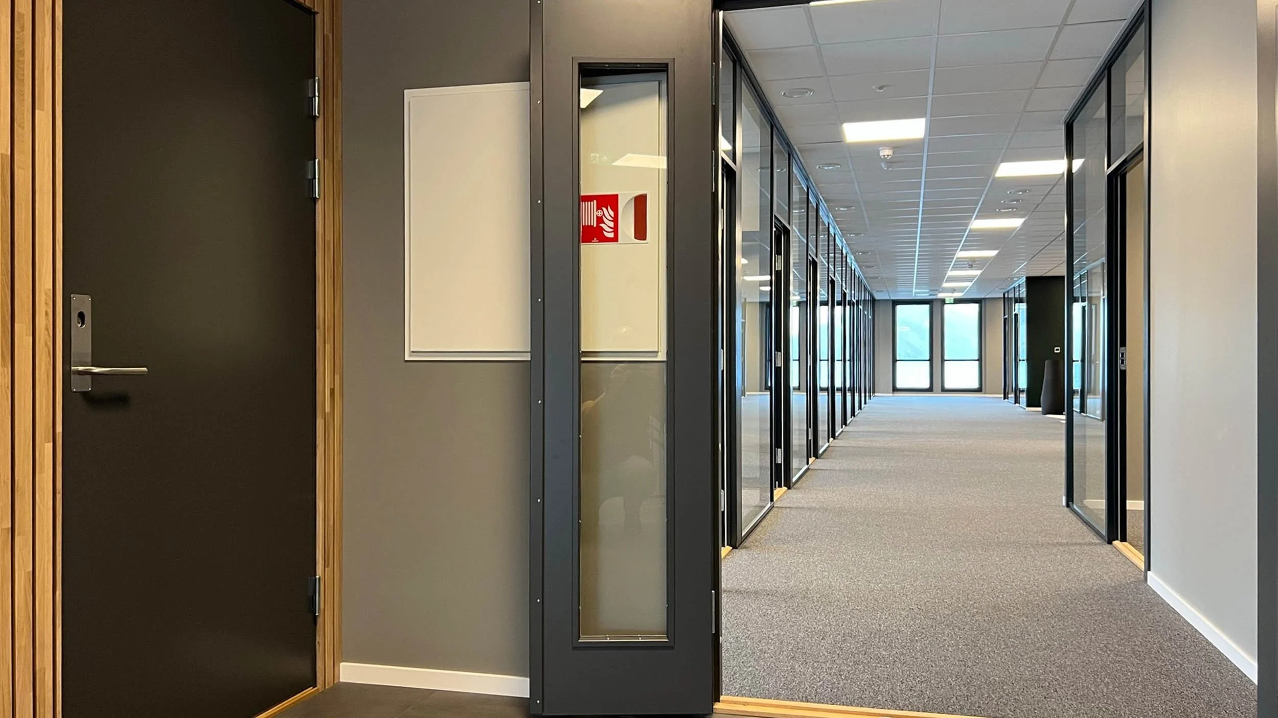 Et moderne kontorlandskap med glassveggdører og kontorer langs en korridor. Det er godt opplyst med taklys og store vinduer i enden.