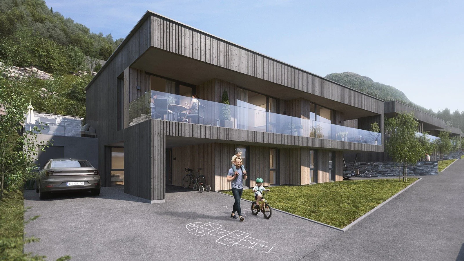 Moderne leilighetsbygg med balkonger, personer som går med barn, parkeringsplass og lekeområde, omgitt av grønt landskap og fjell.