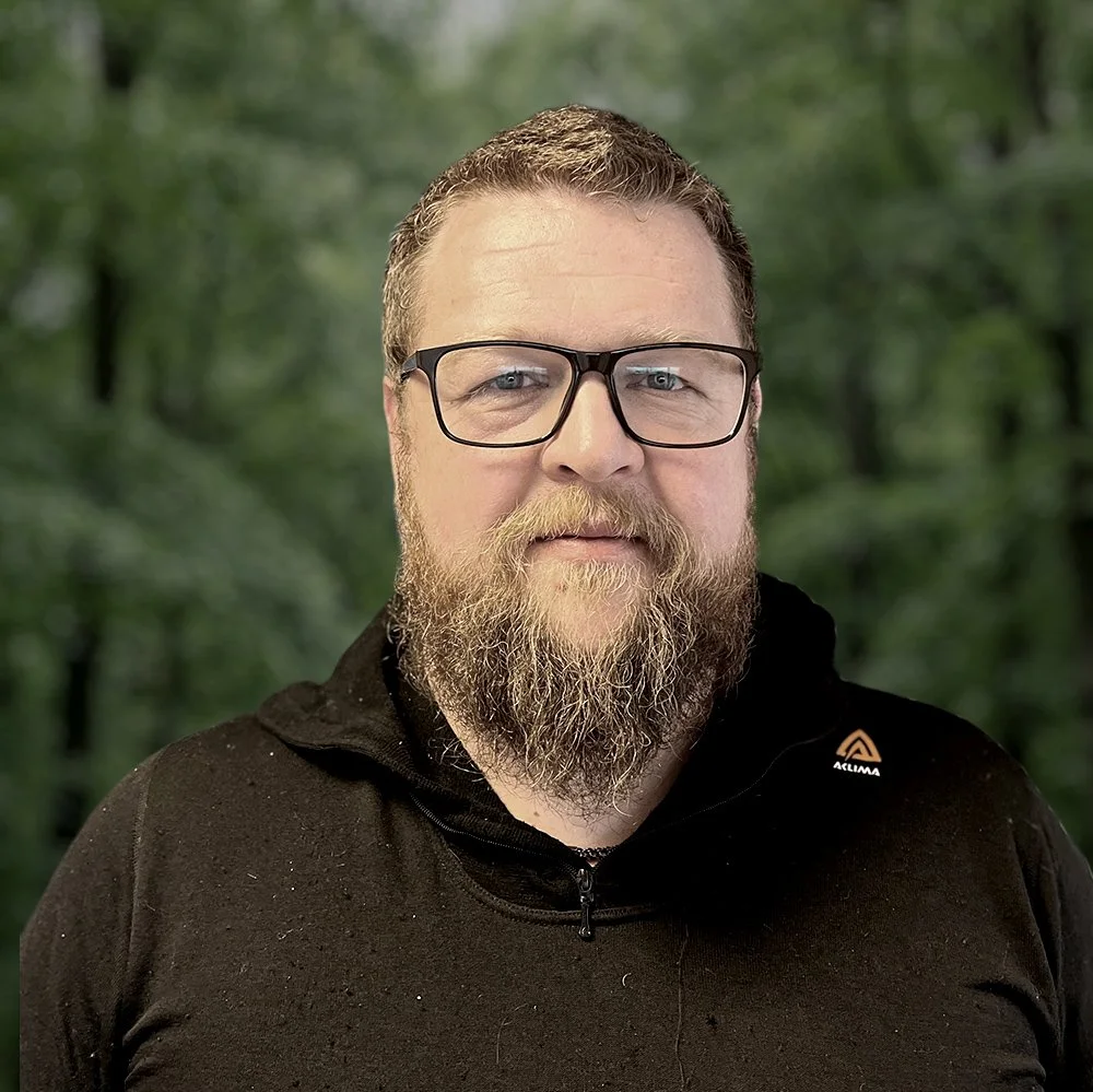 En mann med brunt, krøllet hår og skjegg, iført briller og svart genser, står i en grønn skog.