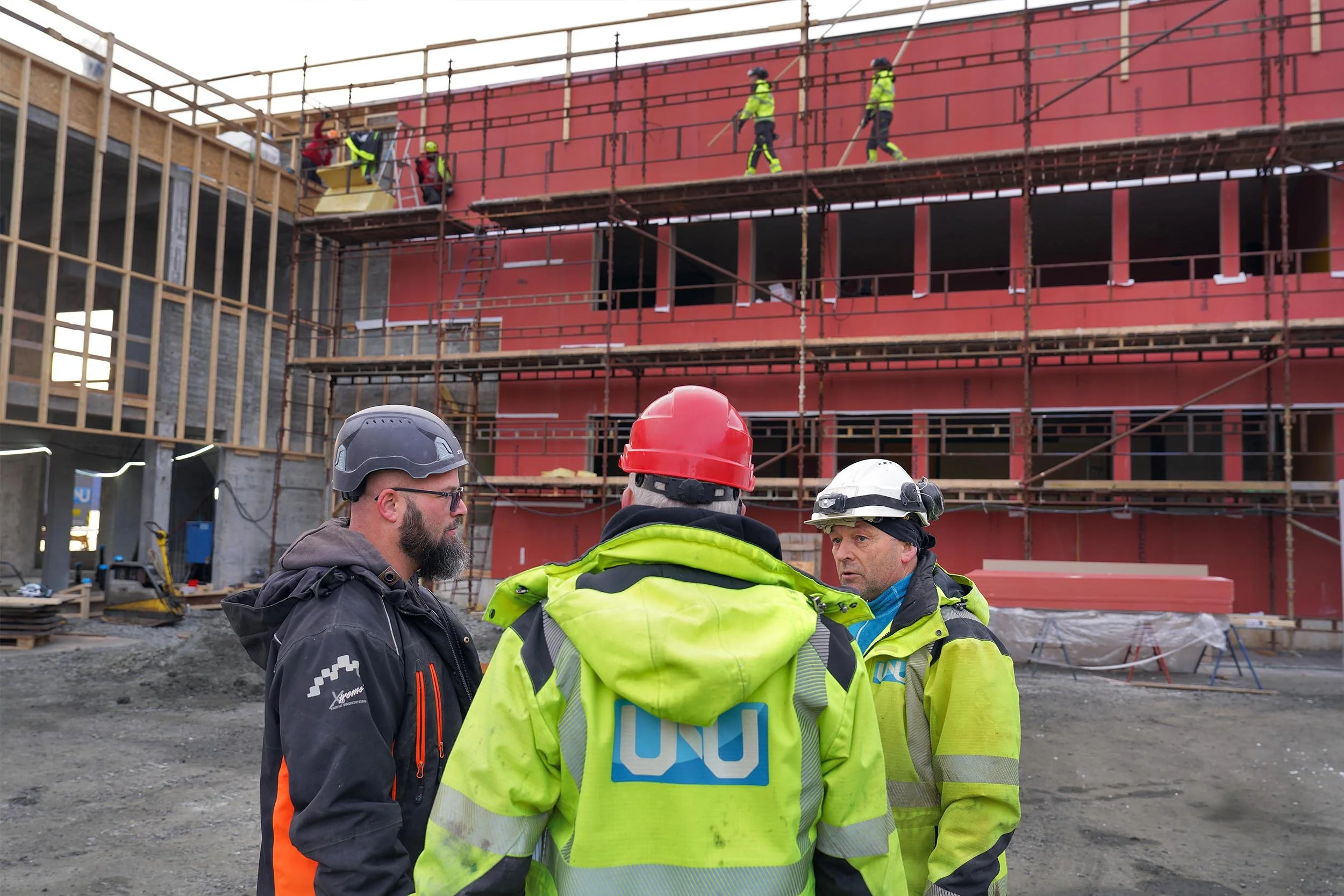 Grupp av byggearbeidere med hjelmer og refleksvester foran en byggeplass, med ansatte som arbeider på et stillas i bakgrunnen.