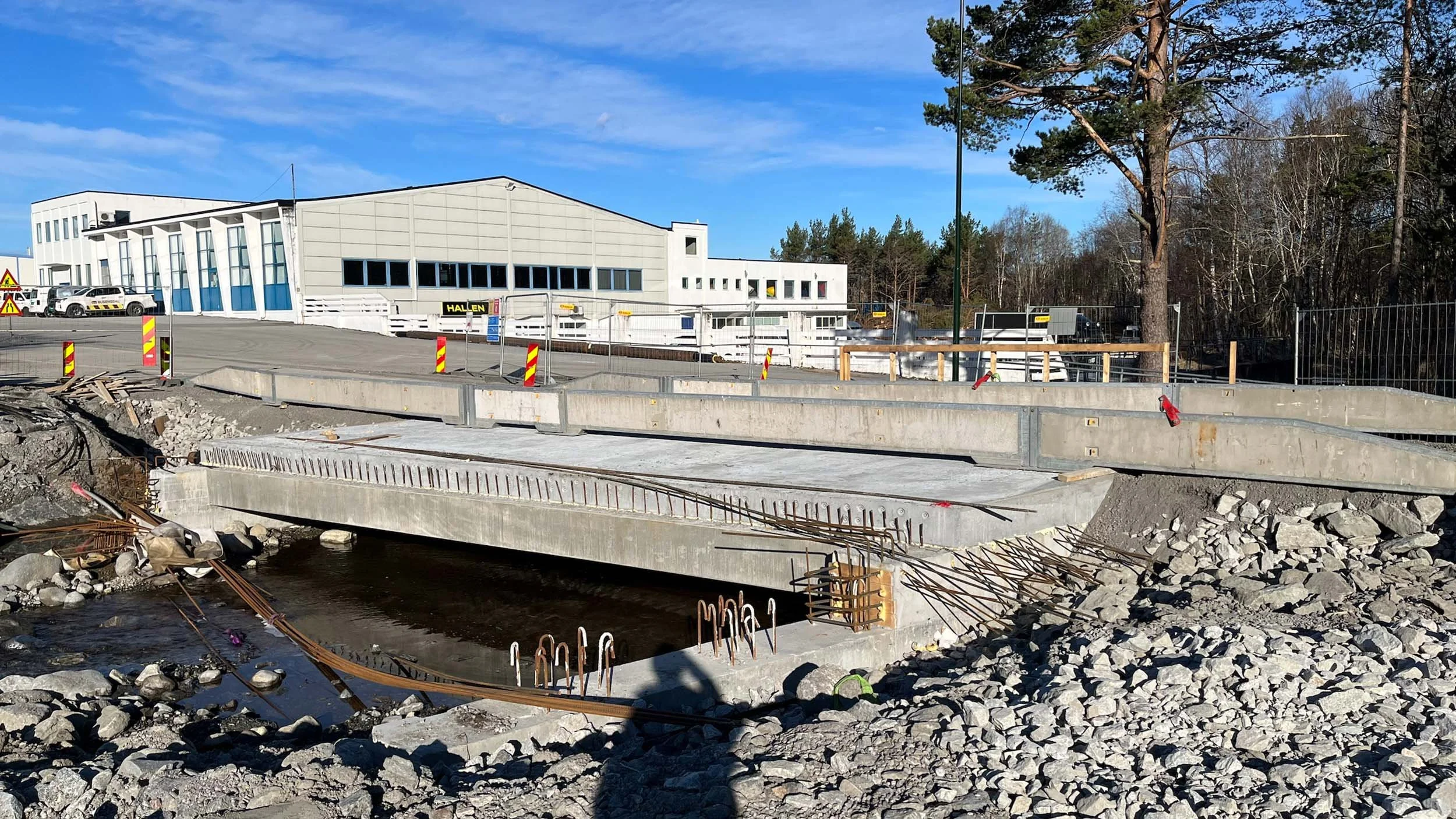 Pågår byggearbeid med en ny bro eller vei på et område med stein og jord, og en stor hvit bygning i bakgrunnen, omgitt av trær.