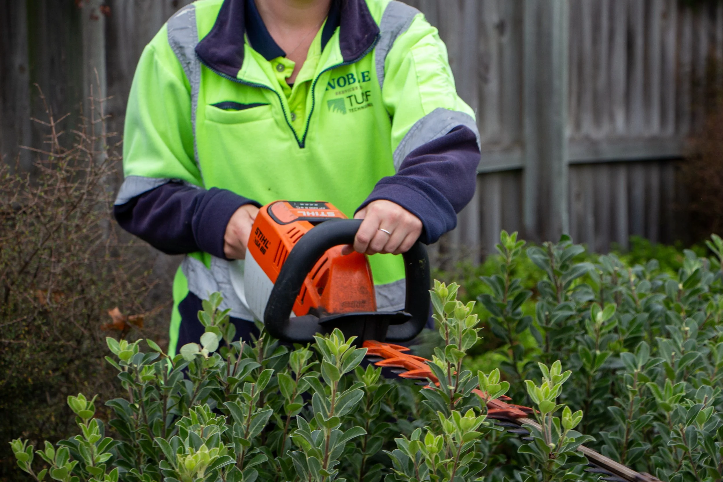 An aerial view of a person wearing a green helmet and orange shirt mowing a lush green lawn with a lawn mower. The lawn is bordered by flower beds with various plants and flowers.