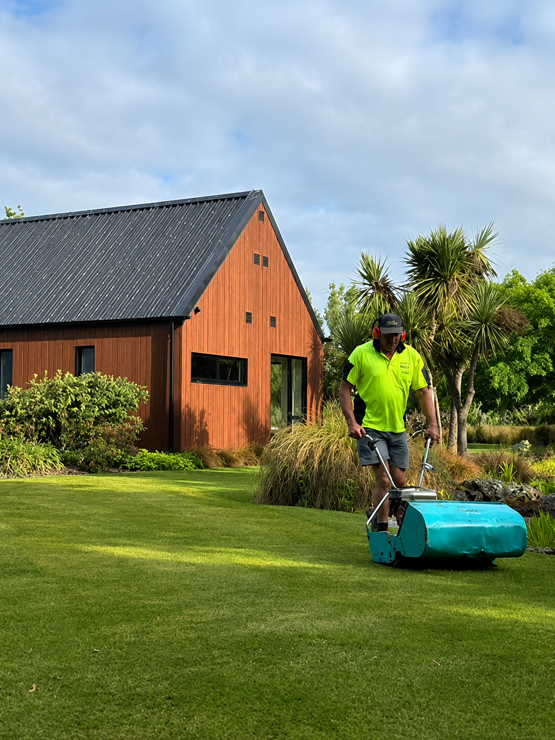 A man in a bright yellow shirt and shorts using a blue lawn roller on a well-manicured lawn in front of a wooden house with a black metal roof, surrounded by trees and greenery under a partly cloudy sky.