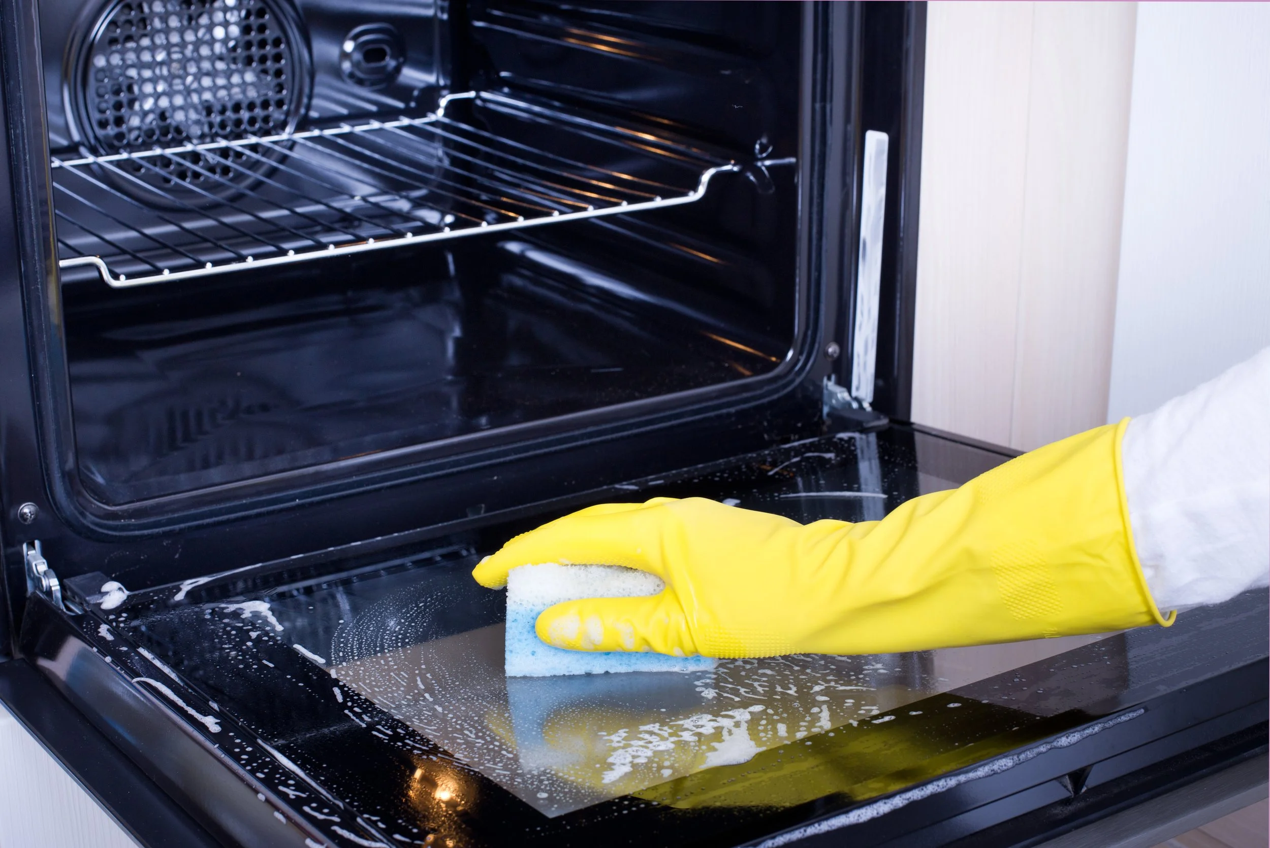 Person wearing yellow rubber gloves cleaning the inside of a black oven with a sponge.