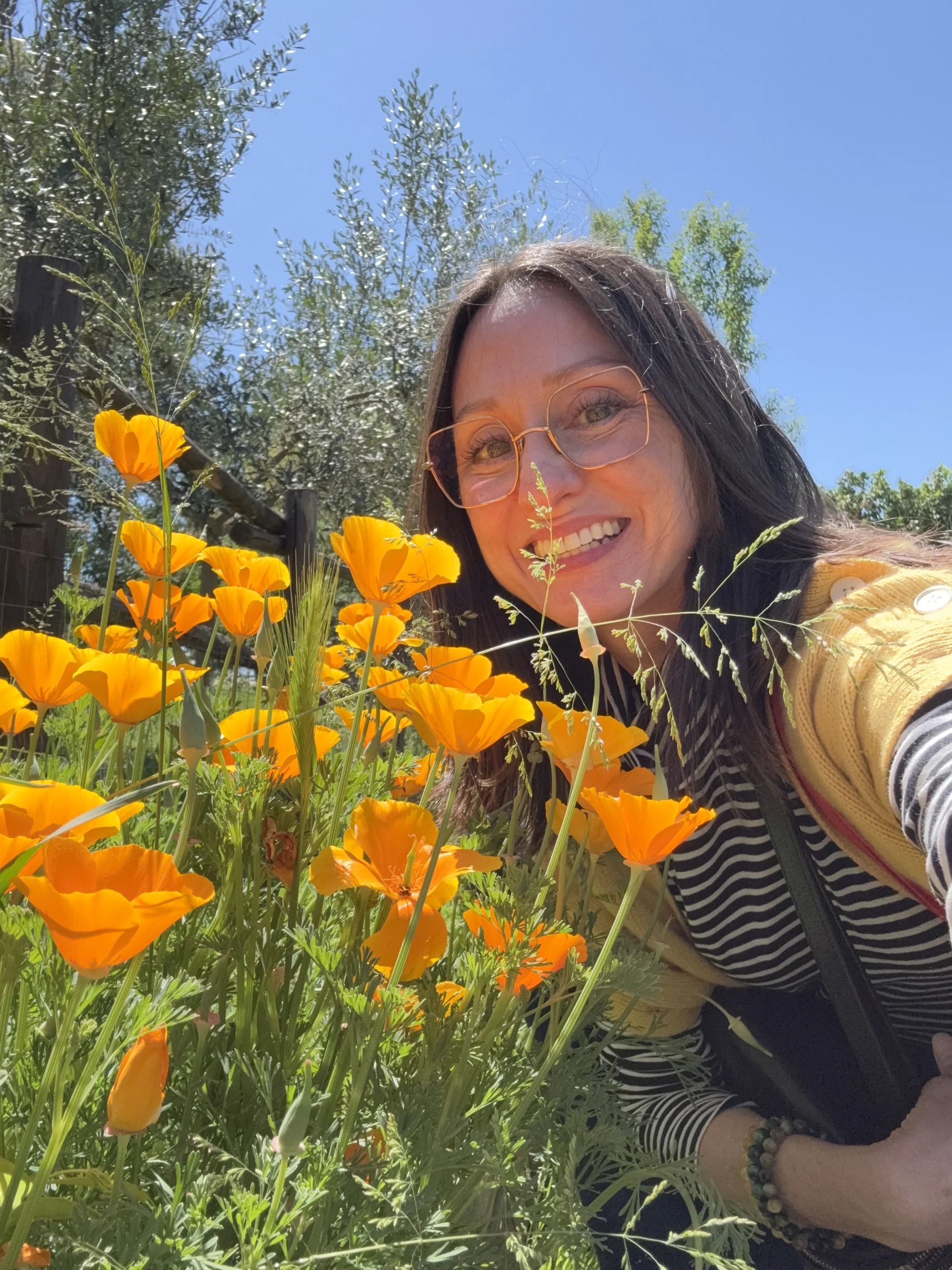 A woman with glasses and a striped shirt smiling outdoors among orange flowers and green foliage on a sunny day.