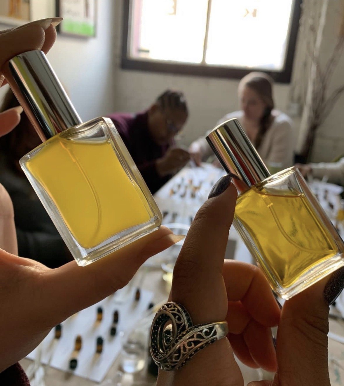 Two hands holding small rectangular glass bottles filled with yellow liquid, with the background showing two women sitting at a table.