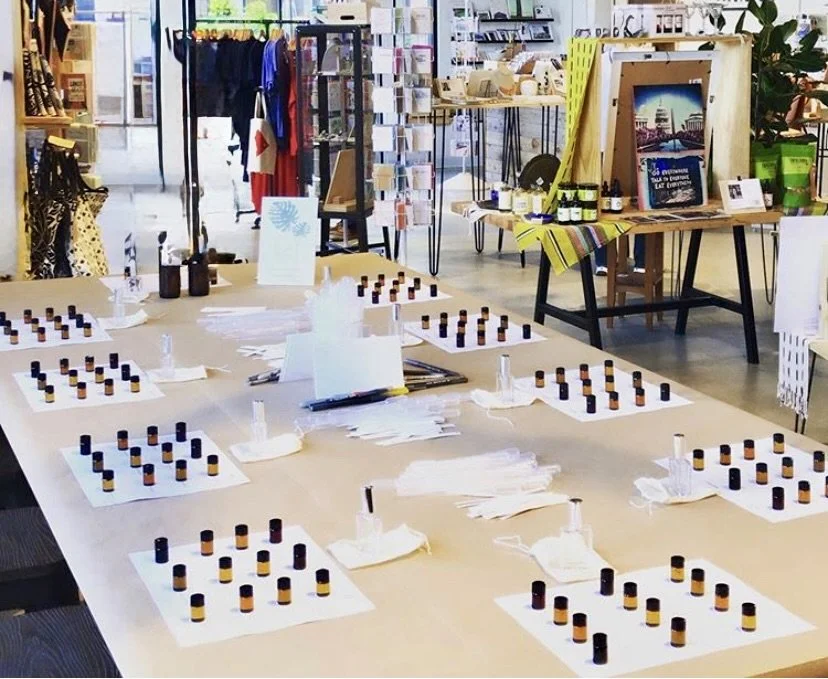 A table in a retail store with multiple blotter paper sheets, each with several small bottles arranged in rows, likely for a scent or essential oil sampling event. Shelves of clothing and display tables are visible in the background.