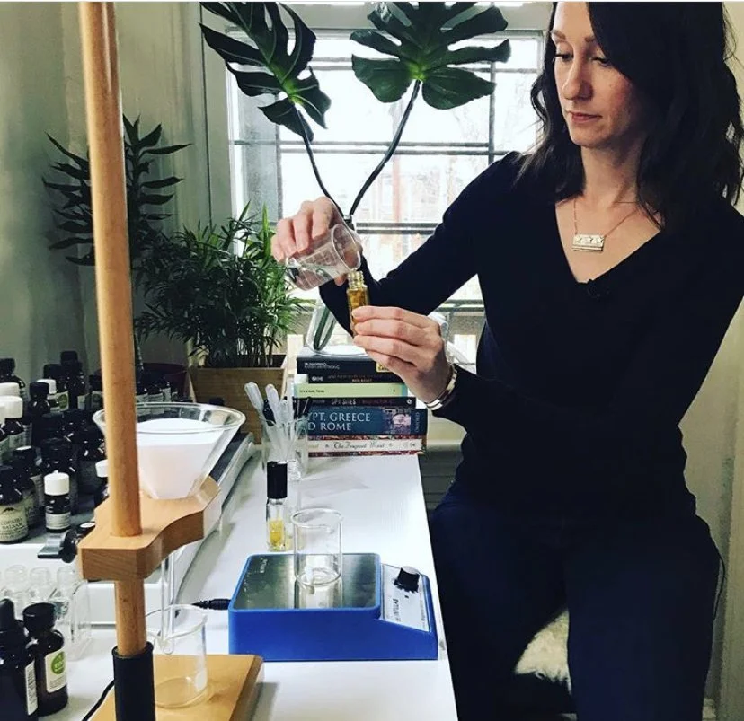 A woman working with essential oils at a desk, holding a glass container with a dropper, surrounded by bottles, books, and plants.