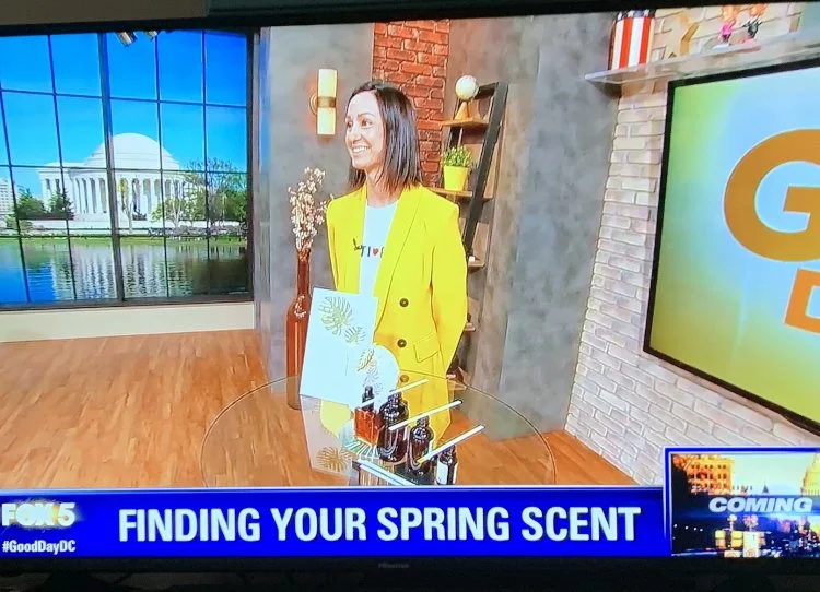 TV studio with a woman in a yellow blazer standing behind a display of spring-scented products, with a large screen showing 'Good Day DC' and a backdrop of the U.S. Capitol building visible through large windows.