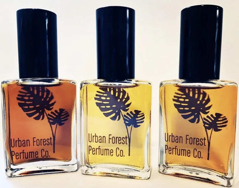 Three bottles of perfume from Urban Forest Perfume Co. with dark blue caps, containing orange, yellow, and orange-colored liquids.