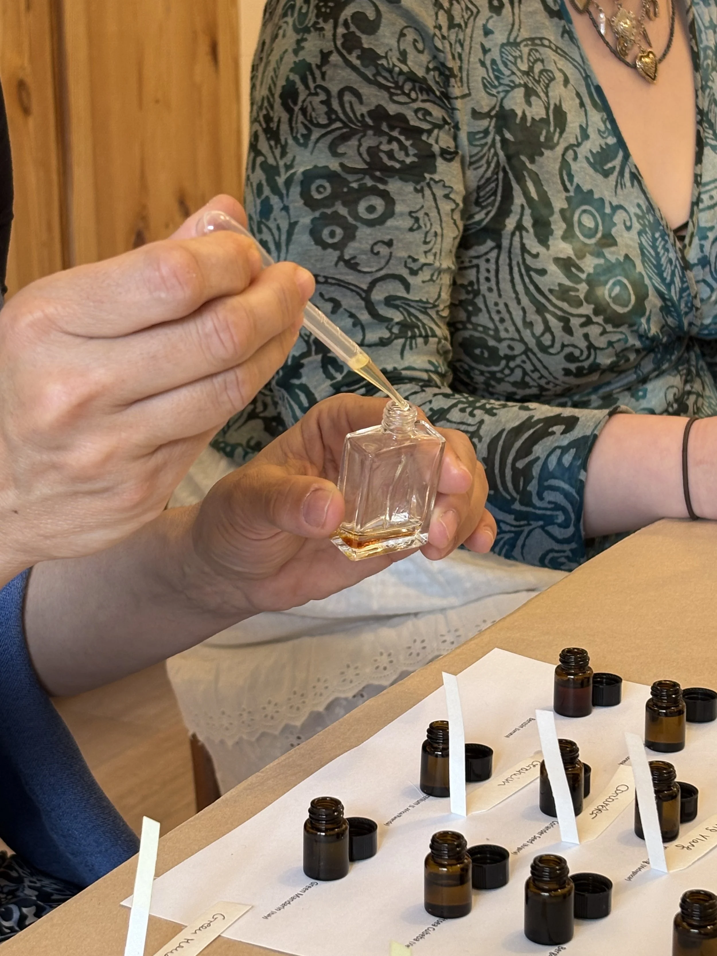 Person using a dropper to transfer liquid into a small glass bottle, with several labeled small bottles and handwritten labels on a table.