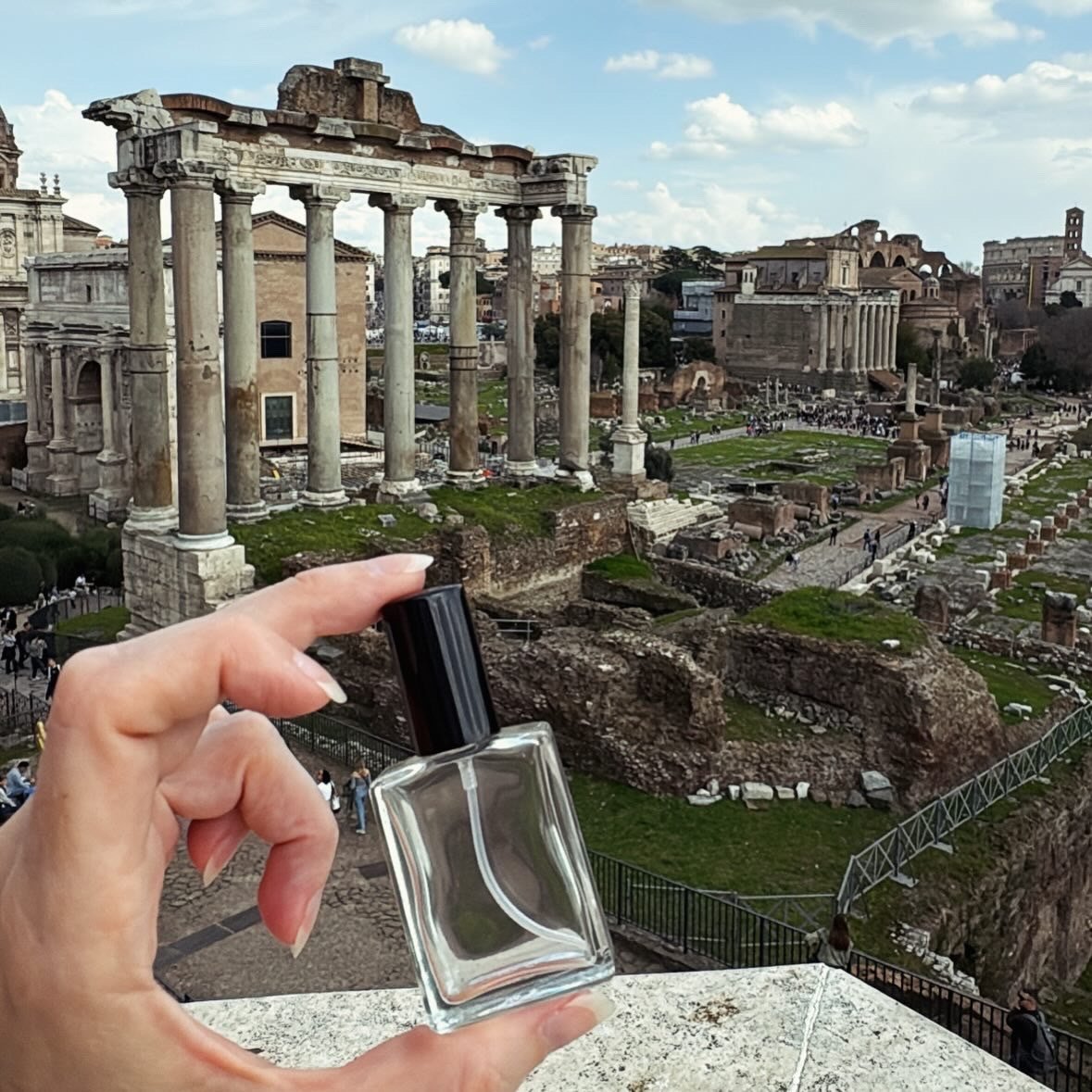 A hand holding a perfume bottle in front of ancient Roman ruins, likely in Rome, Italy, with a cityscape in the background under a partly cloudy sky.