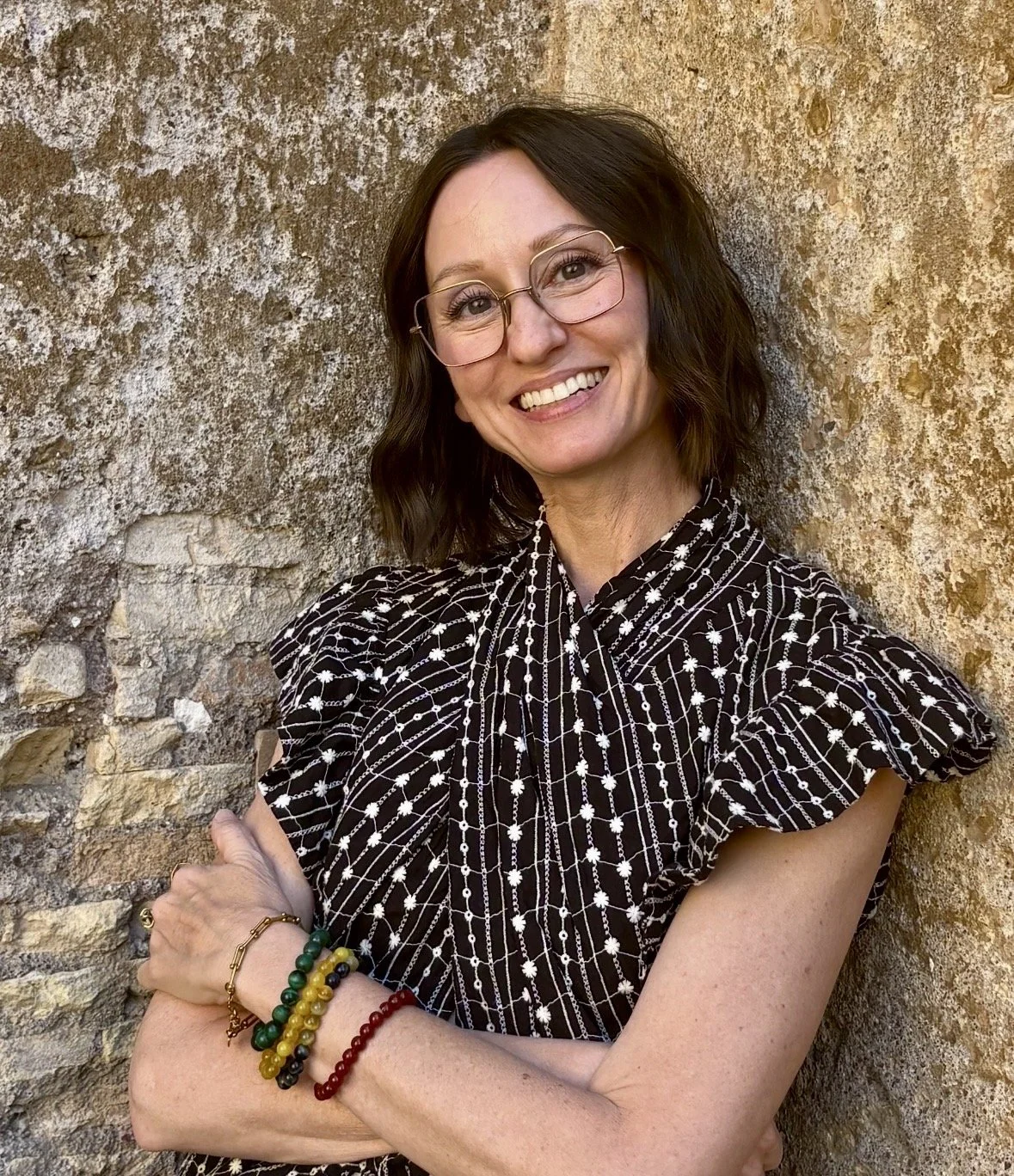 A woman with shoulder-length dark hair, wearing glasses, smiling, standing against a textured stone wall. She is dressed in a black and white patterned top with puffed sleeves and accessorized with colorful bracelets on her left wrist.