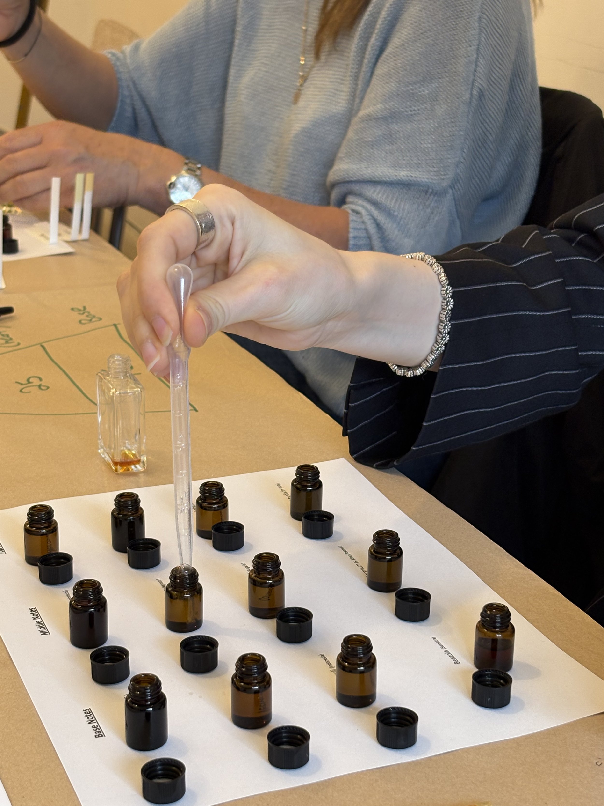 People examining bottles of essential oils or fragrances arranged on a white sheet with labeled sections, on a wooden table.