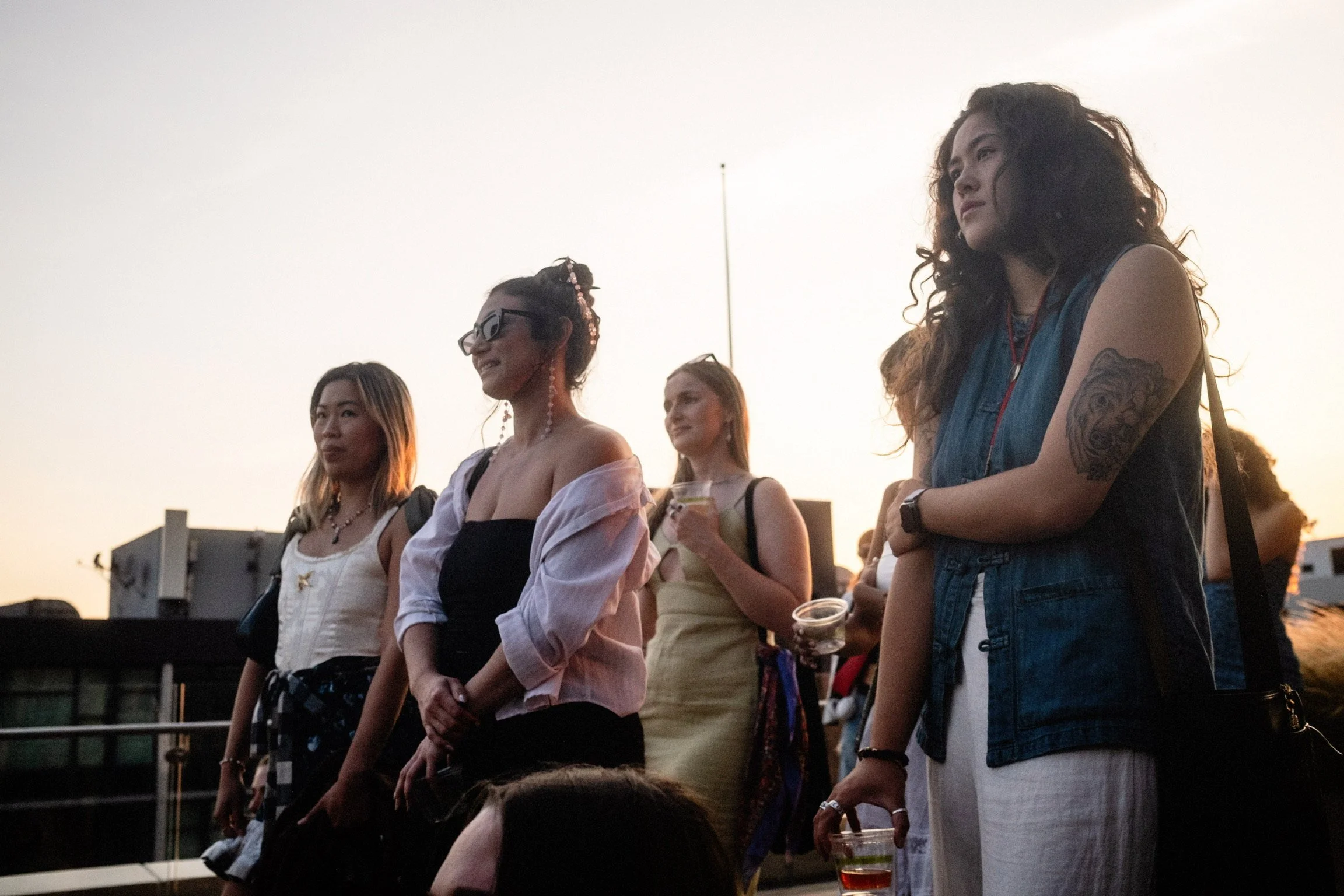 A group of women standing outdoors during sunset, some holding drinks, with buildings in the background.