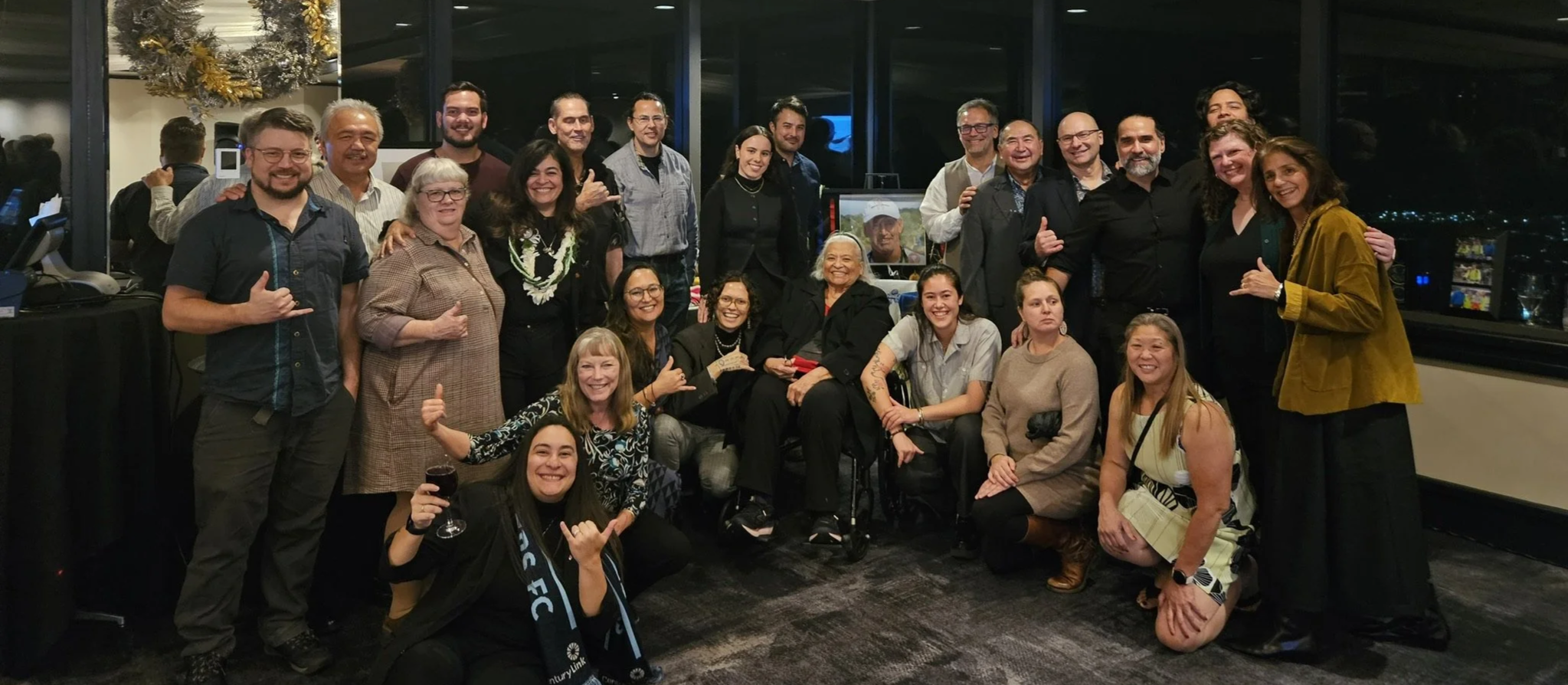 A large group of people gathered together at an indoor event, posing for a group photo. They are smiling and appear to be celebrating. The setting has large windows with a night view outside, and there's a Christmas wreath hanging on the wall.
