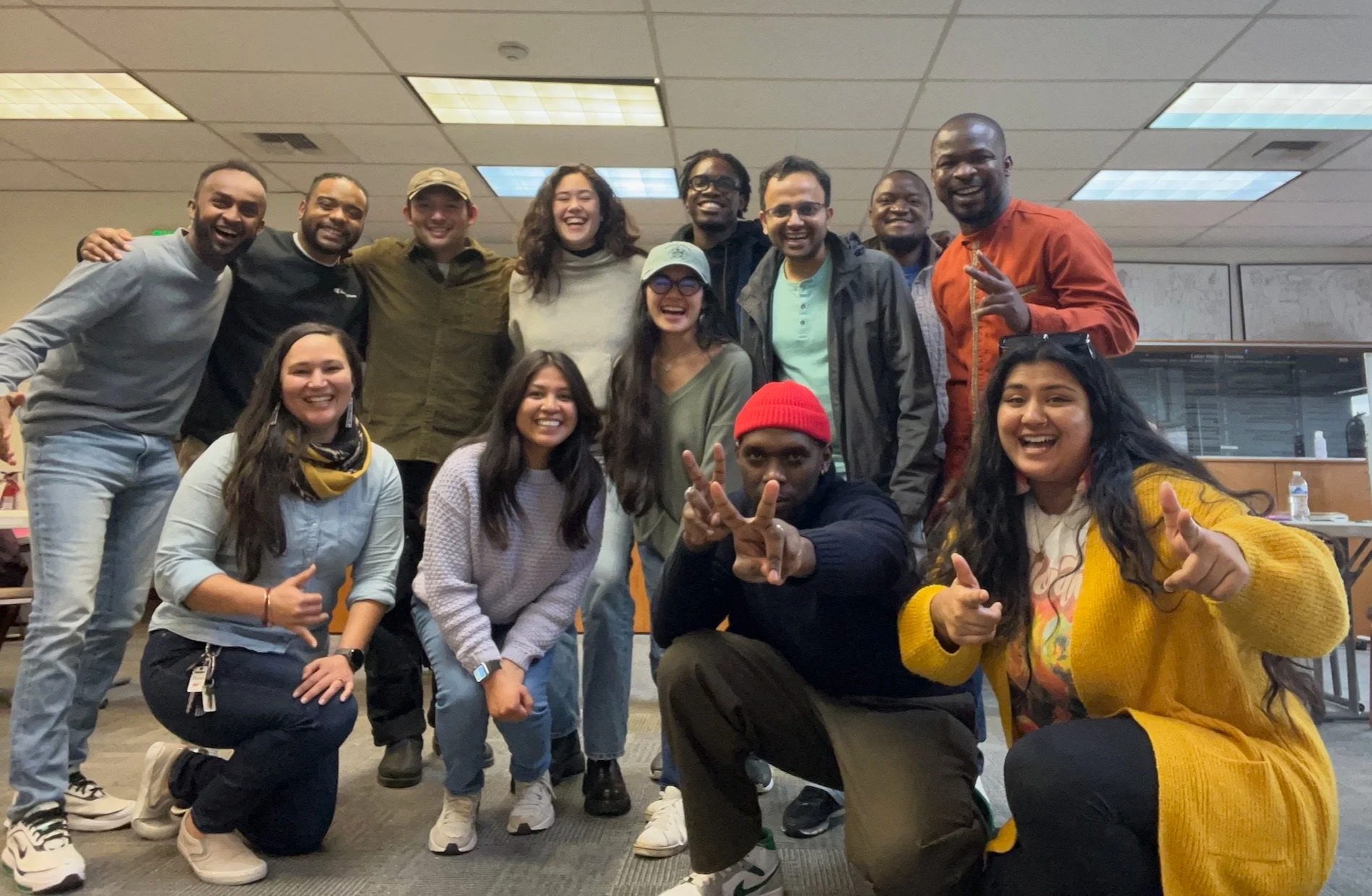 Group of diverse young people in an office, smiling and making peace signs, posing together for a photo.