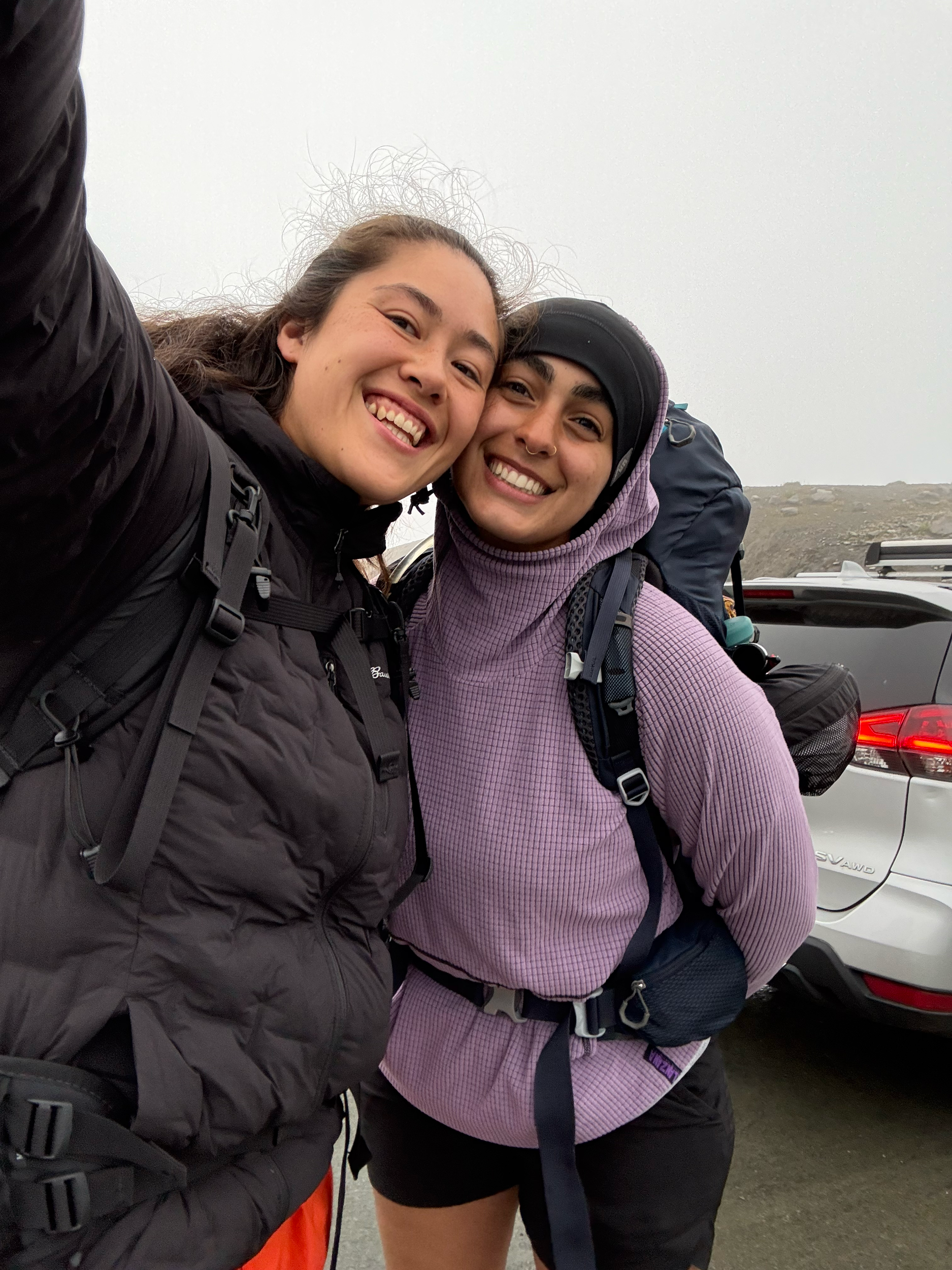 Two smiling women with backpacks taking a selfie outdoors on a foggy day. One woman has curly hair and is wearing a black jacket, and the other has straight hair, wearing a purple hoodie and a black cap.