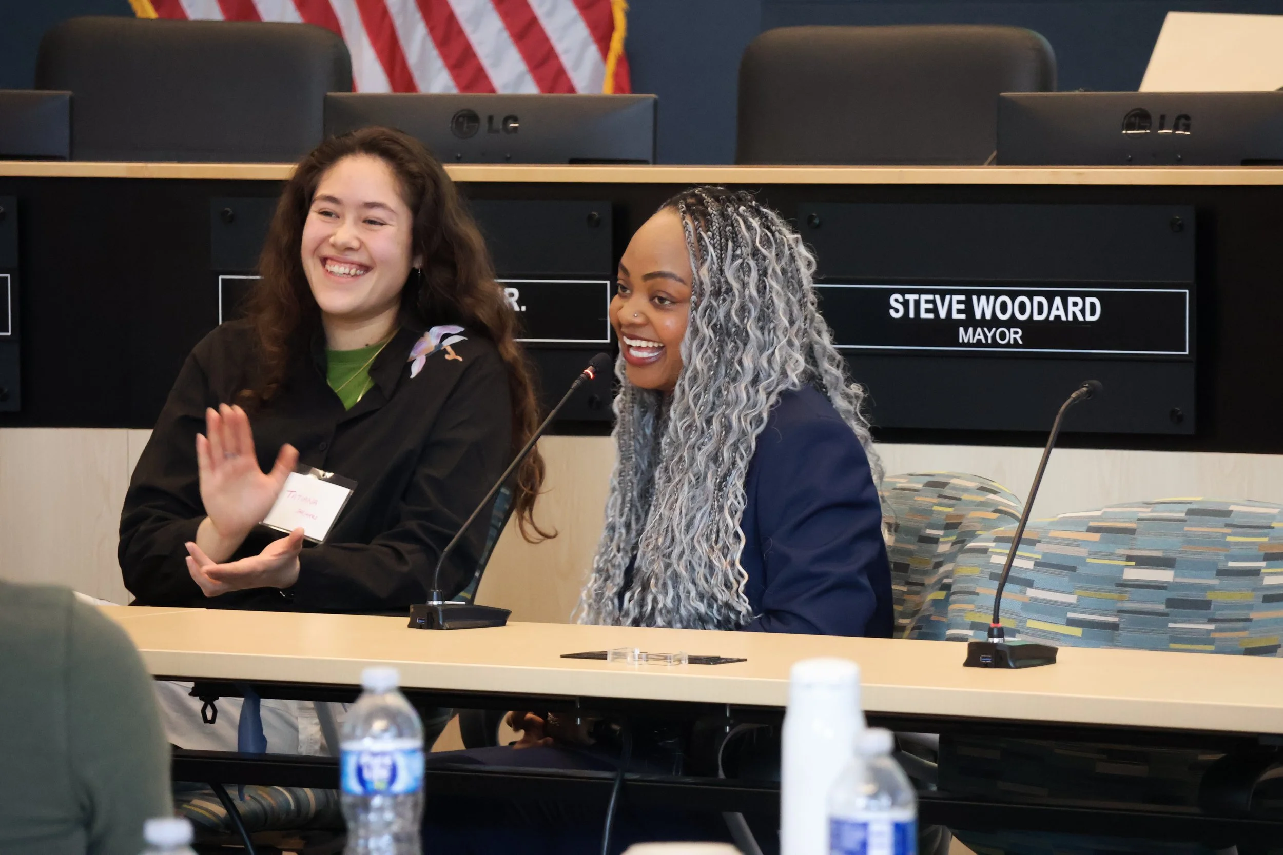 Two women sitting at a table, smiling and laughing, in a meeting room with nameplates and microphones. One woman has long, curly brown hair, and the other has long, blonde and gray braided hair. A sign for Mayor Steve Woodard is visible in the backgr