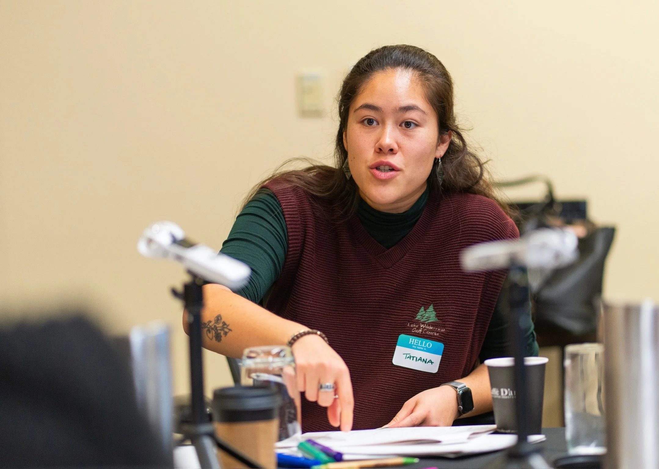 Tatiana in action at a community assembly. She is a young woman with brown curly hair, wearing a green and maroon shirt, is sitting at a table with craft supplies in front of her.