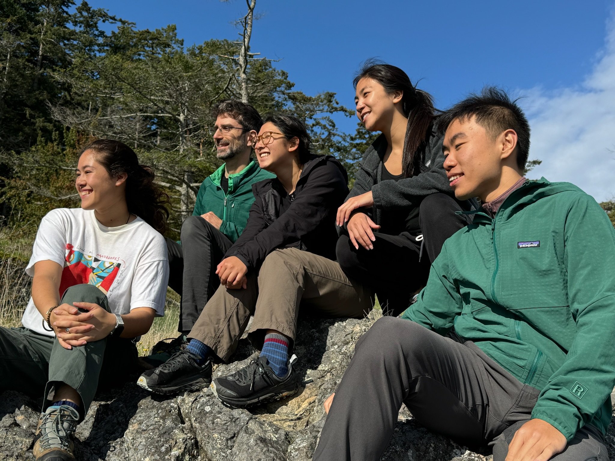 Five people sitting outdoors on rocky ground, smiling and looking to the right, with trees and blue sky in the background.