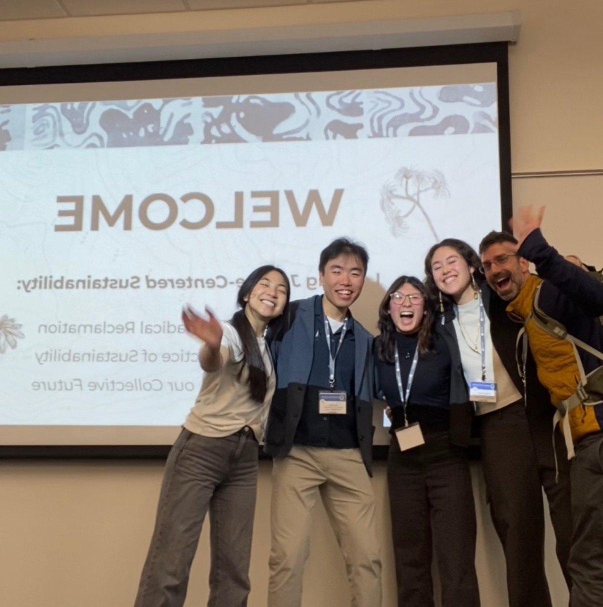 Group of five people at a conference from the Campus Sustainability Fund, smiling and posing in front of a presentation screen that says "WELCOME" and details about a research sustainability event.