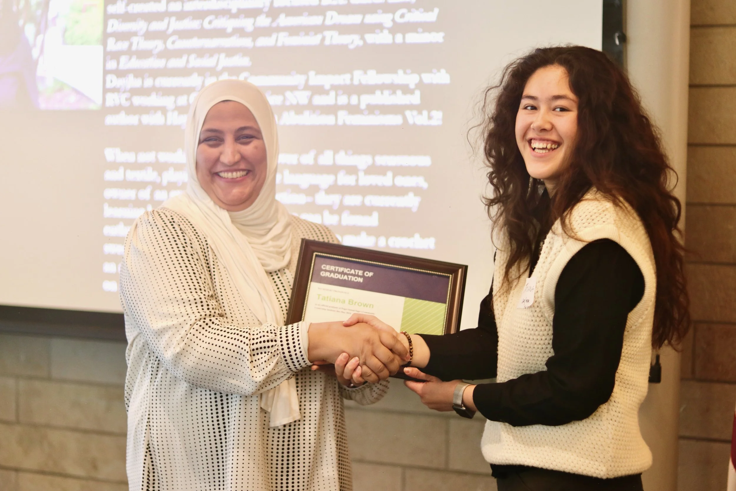 Two women are shaking hands in front of a presentation screen. One woman is wearing a white hijab and patterned shirt, and is holding a framed certificate. The other woman has long curly hair and is wearing a cream-colored vest over dark clothing, sm