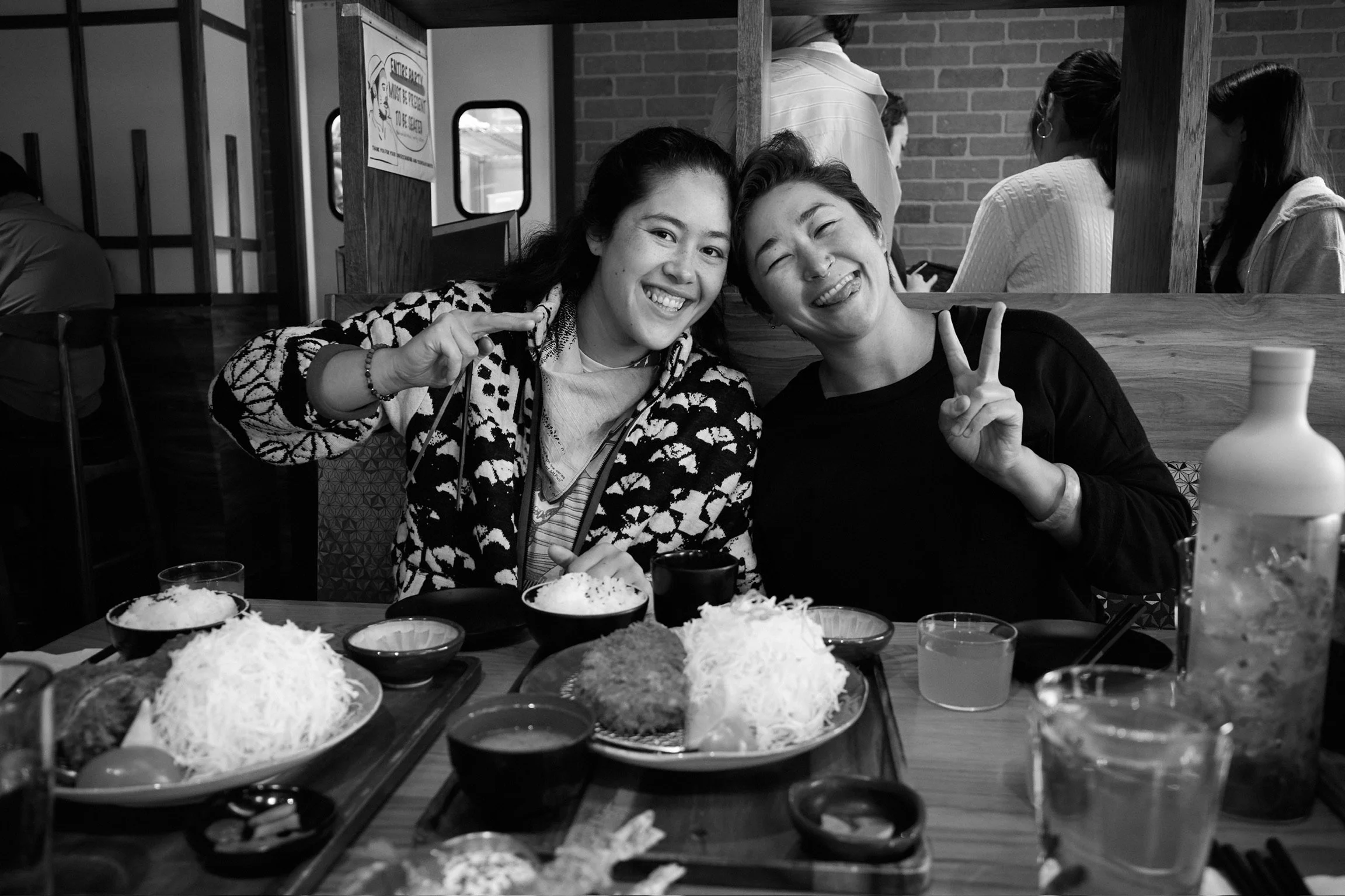Two young women smiling and making peace signs while sitting at a table filled with various dishes in a restaurant environment.