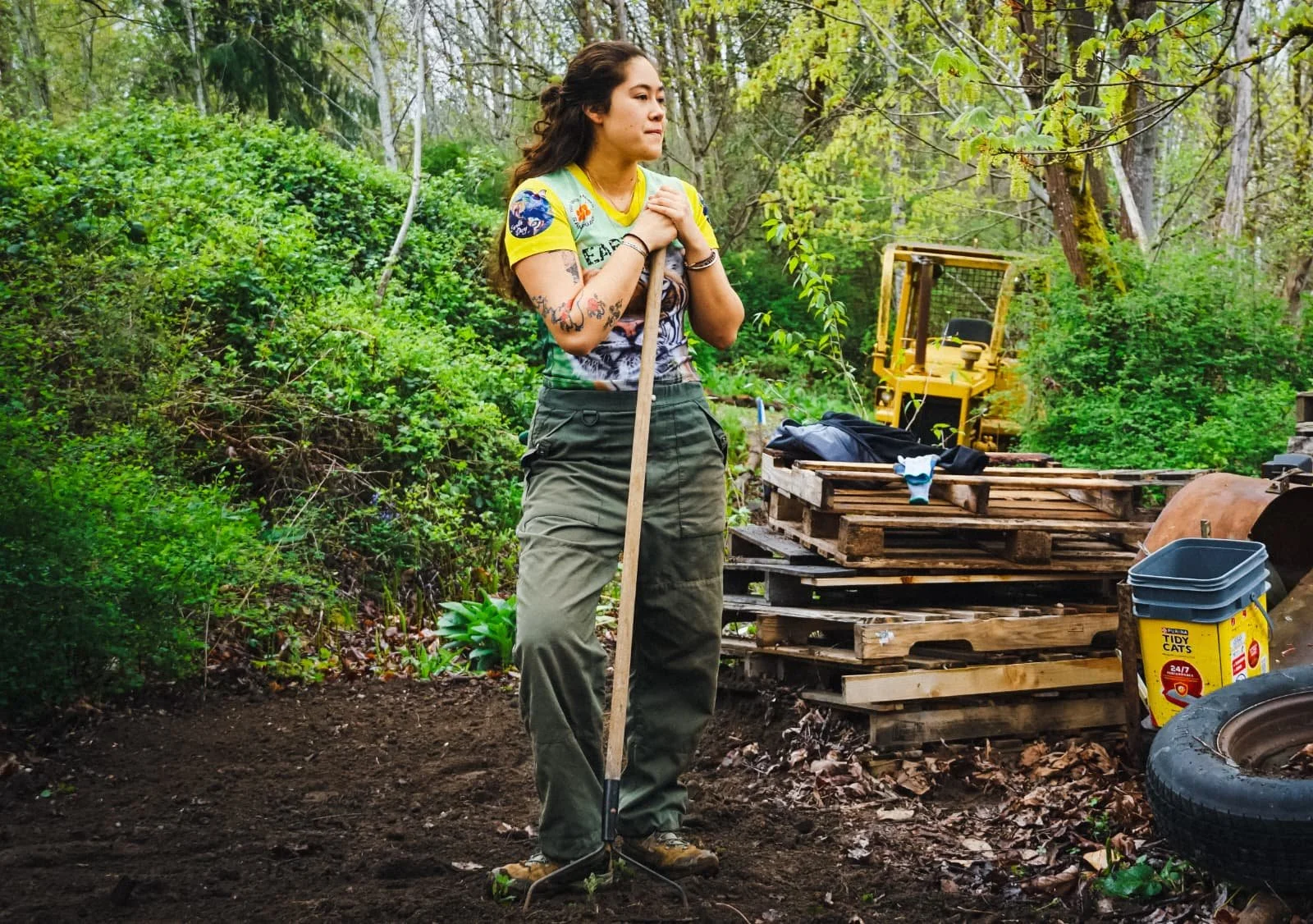 A woman standing outdoors in a forested area, holding a gardening tool, with gardening supplies and tools around her, including a pallet, a bucket, and a tire.