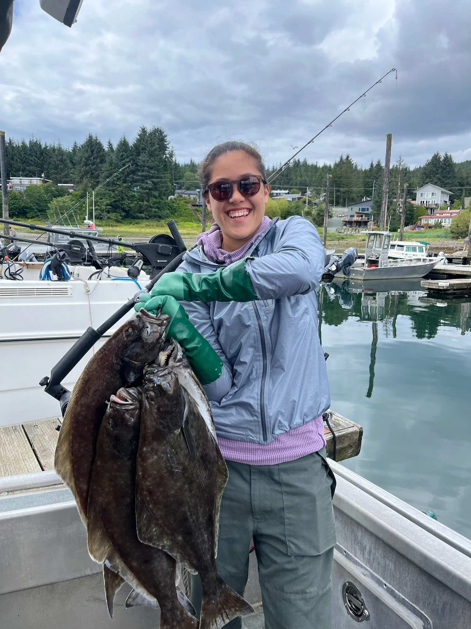 A person wearing sunglasses and a gray jacket holding a large fish on a boat docked in a marina with houses and trees in the background.