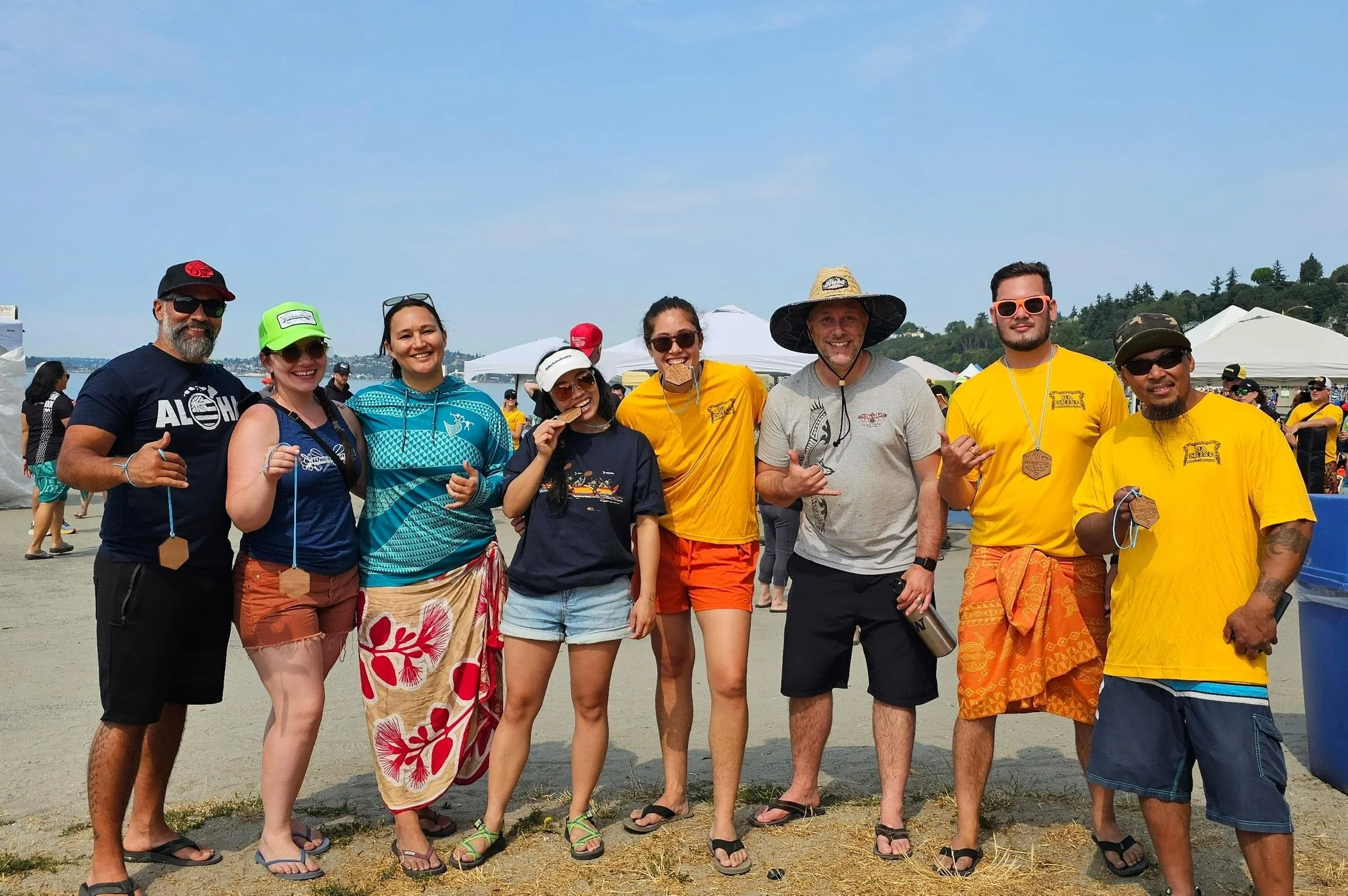 Group of eight people posing outdoors at a festival or event, with tents and a body of water in the background. They are dressed in casual, summery clothing, some wearing sunglasses, hats, and leis, smiling and standing close together.
