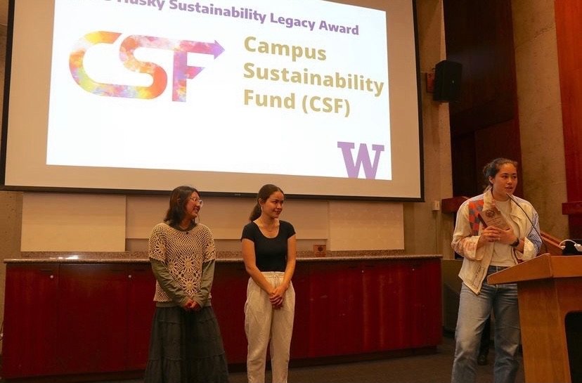 Three women standing in front of a large screen with a presentation slide. The slide displays the text 'Husky Sustainability Legacy Award,' 'CSF,' 'Campus Sustainability Fund (CSF),' and the University of Washington logo.