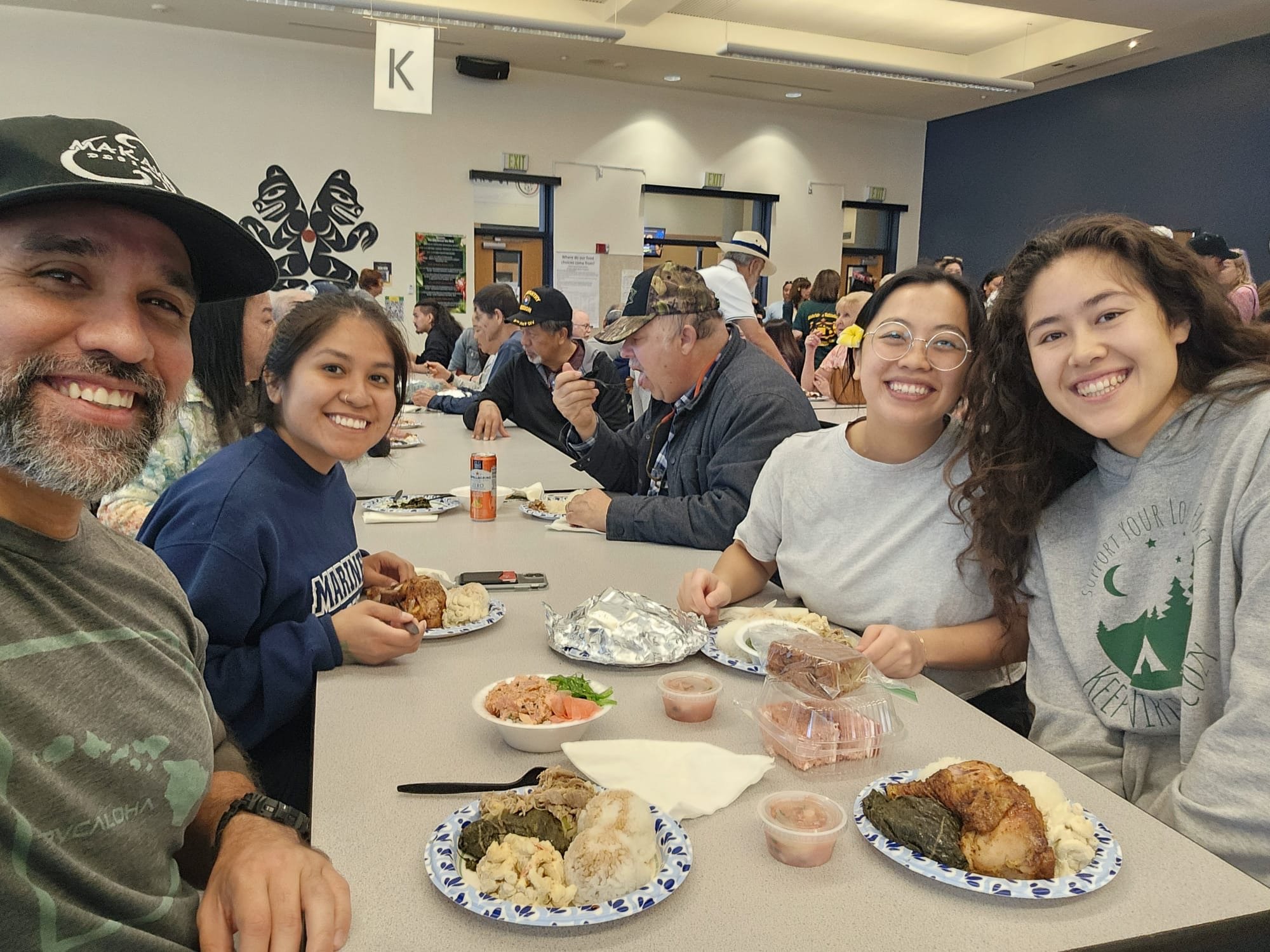 People sitting at a long table in a cafeteria eating traditional Hawaiian food such as spam musubi, laulau, and poke, with some holding plates and some with food in front of them.