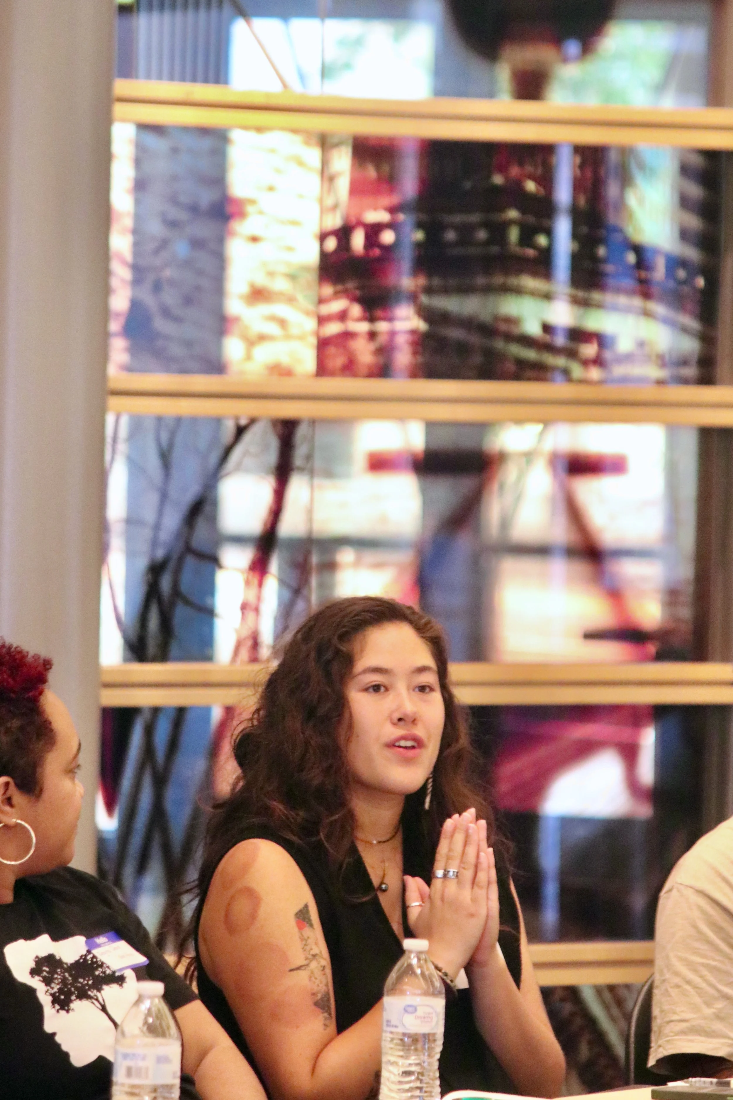 Young woman with dark curly hair and tattoos on her arms, with hands in prayer position, sitting at a table during a group gathering or event.