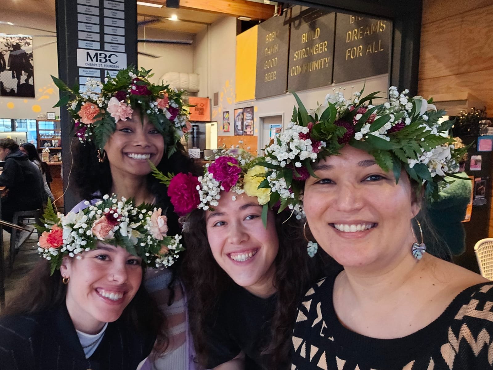 Four women wearing flower crowns, smiling and posing for a photo inside a restaurant or café.