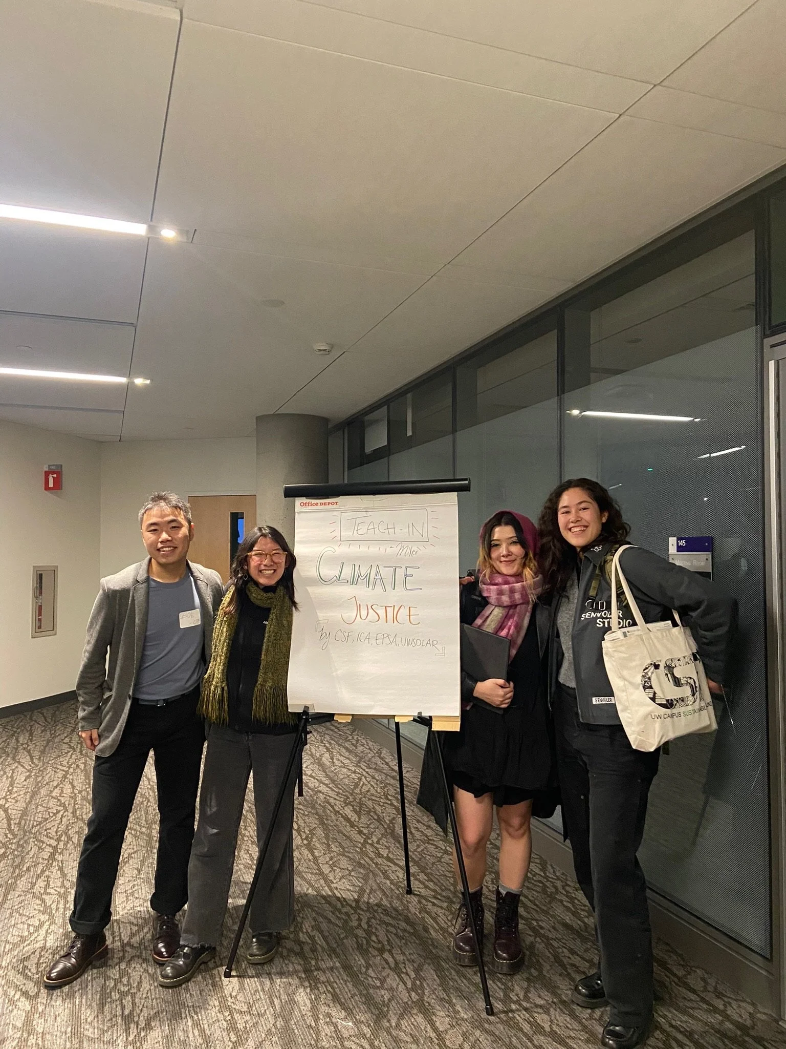 A group of five diverse people standing together indoors near a large sign on an easel that says 'Teach-In Climate Justice' with additional smaller text.