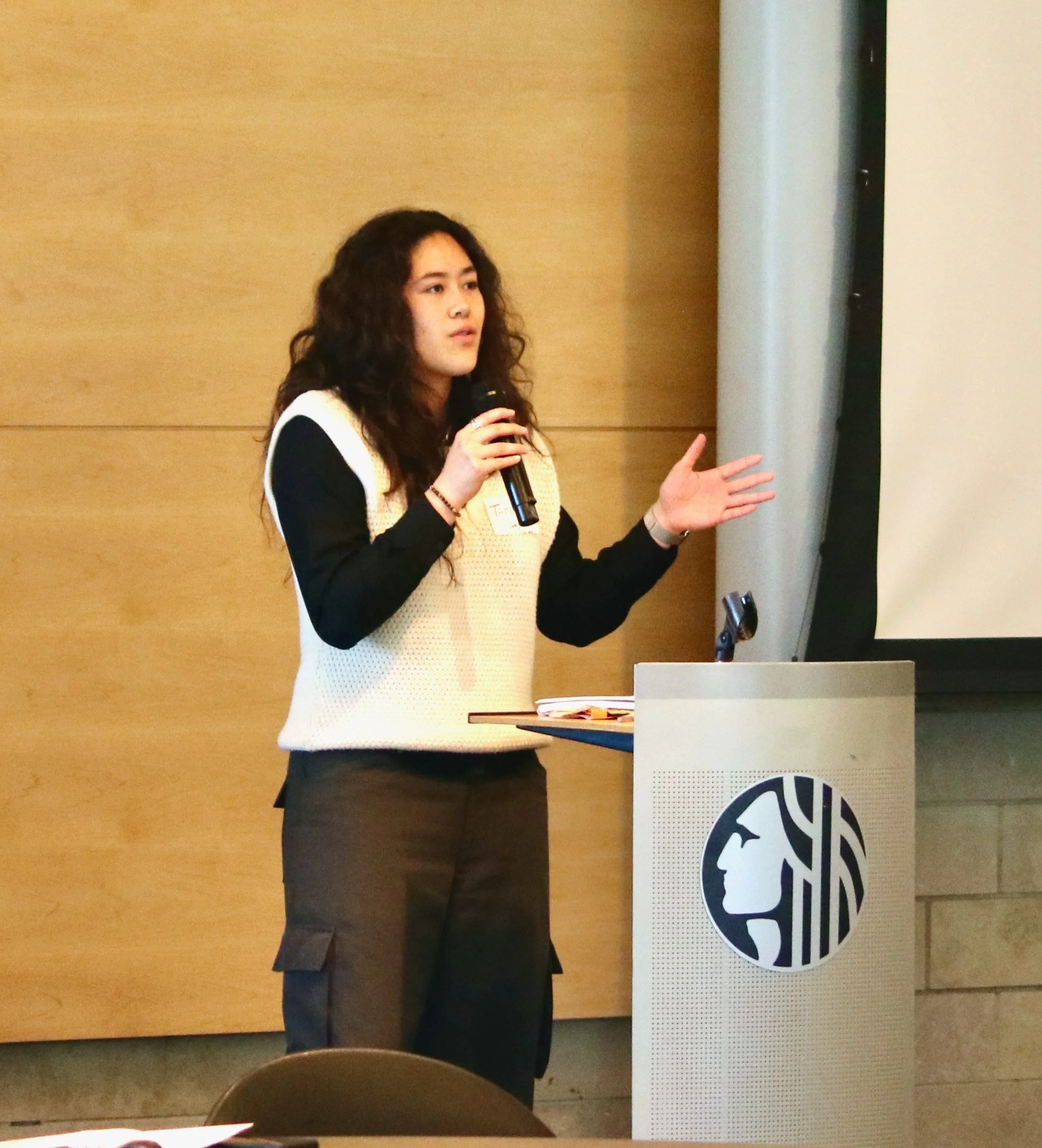 A woman with long curly hair holding a microphone and presenting next to a large screen. She is gesturing with her left hand during her speech. The background is a wood-paneled wall with a proctor logo on a screen or sign.
