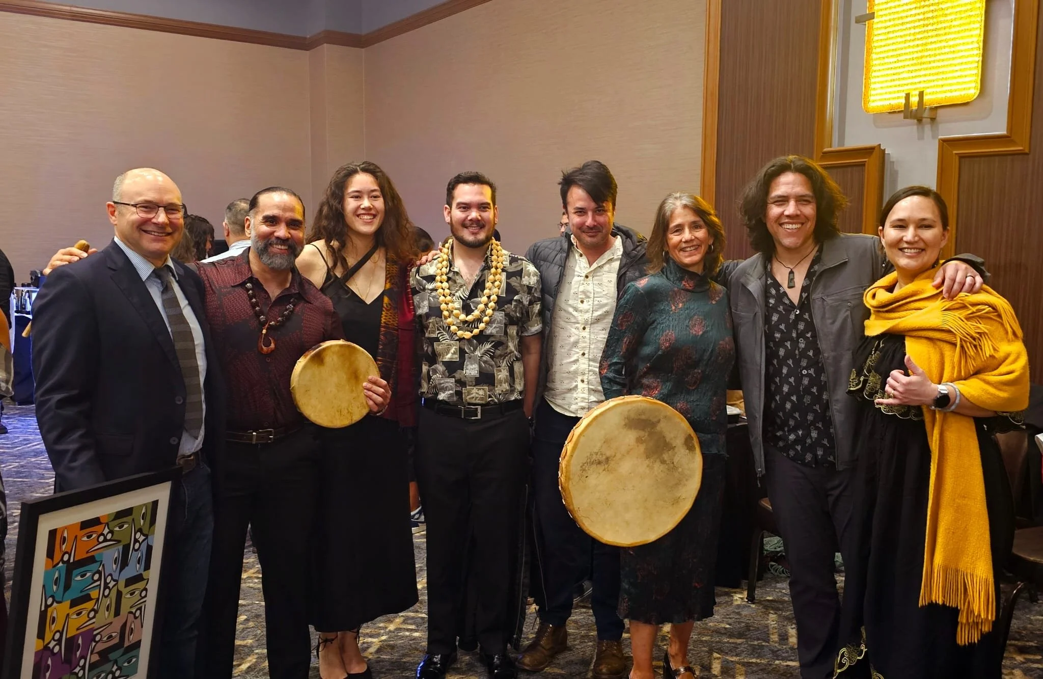 A group of eight diverse people smiling and standing together in an indoor event space, with some holding drums and others wearing traditional or festive clothing.