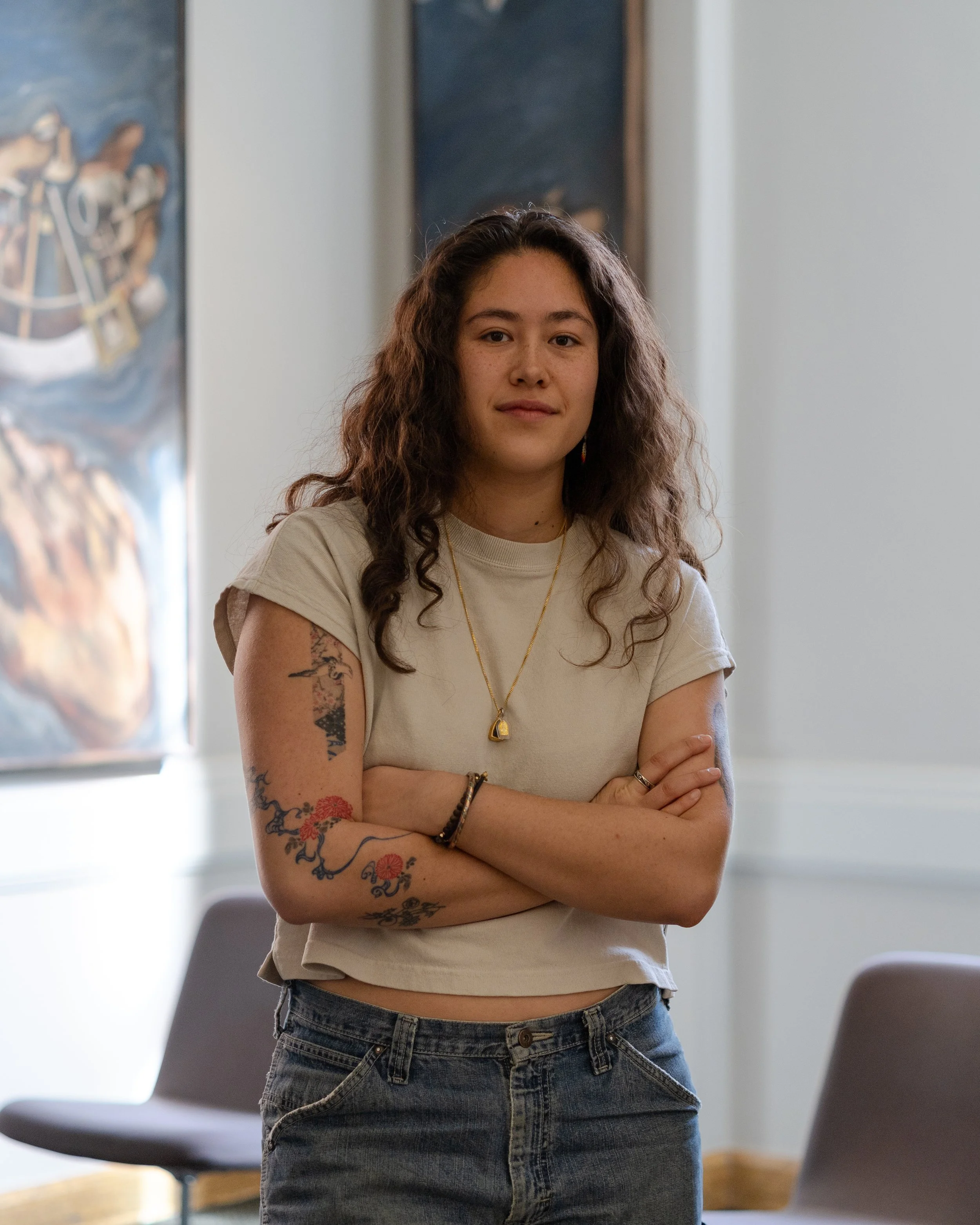 A woman with curly brown hair and tattoos on her arms standing with her arms crossed in a room with chairs and windows in the background.