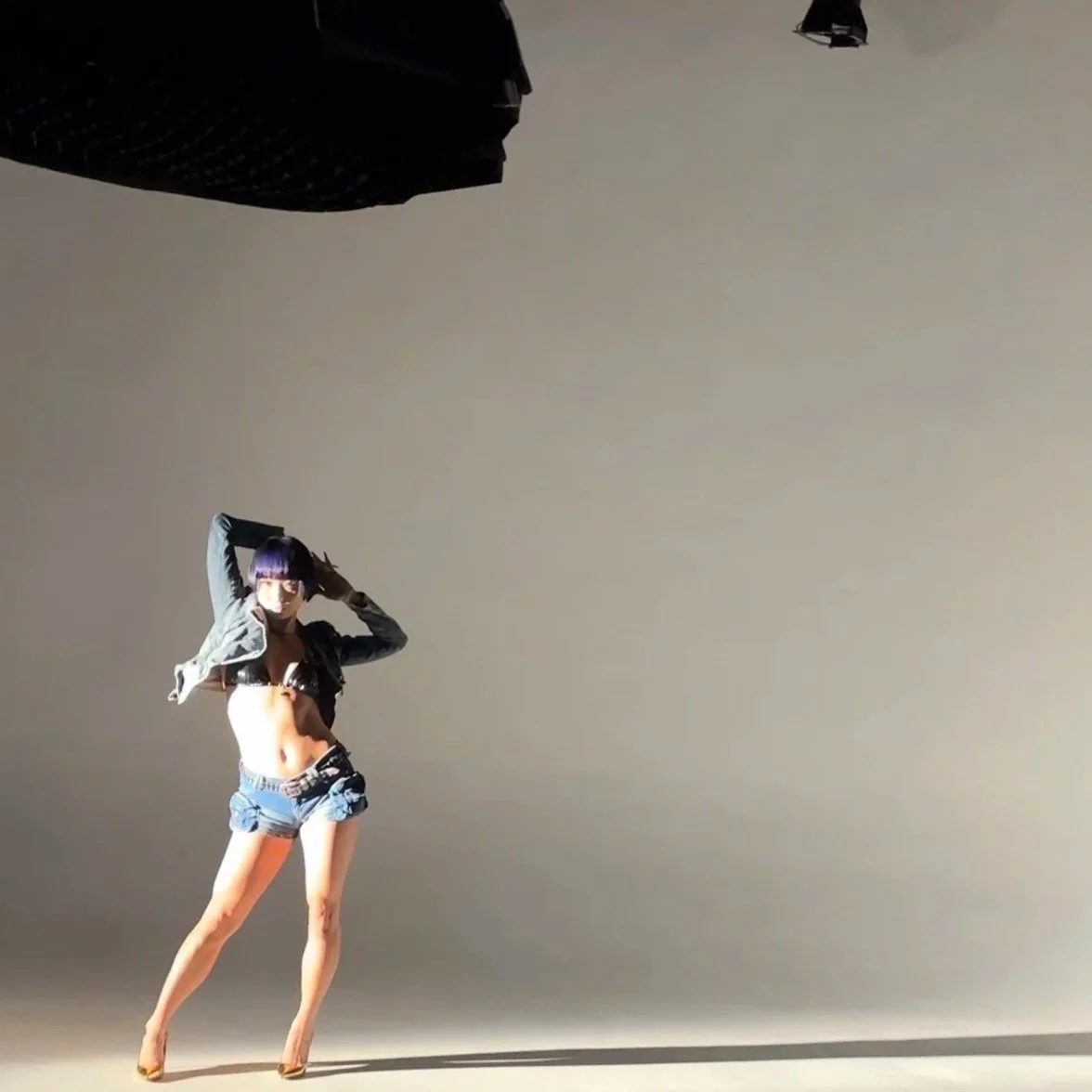 A woman with purple hair wearing a face mask, black crop top, denim shorts, and a jacket standing on a plain background, holding her hair with both hands.
