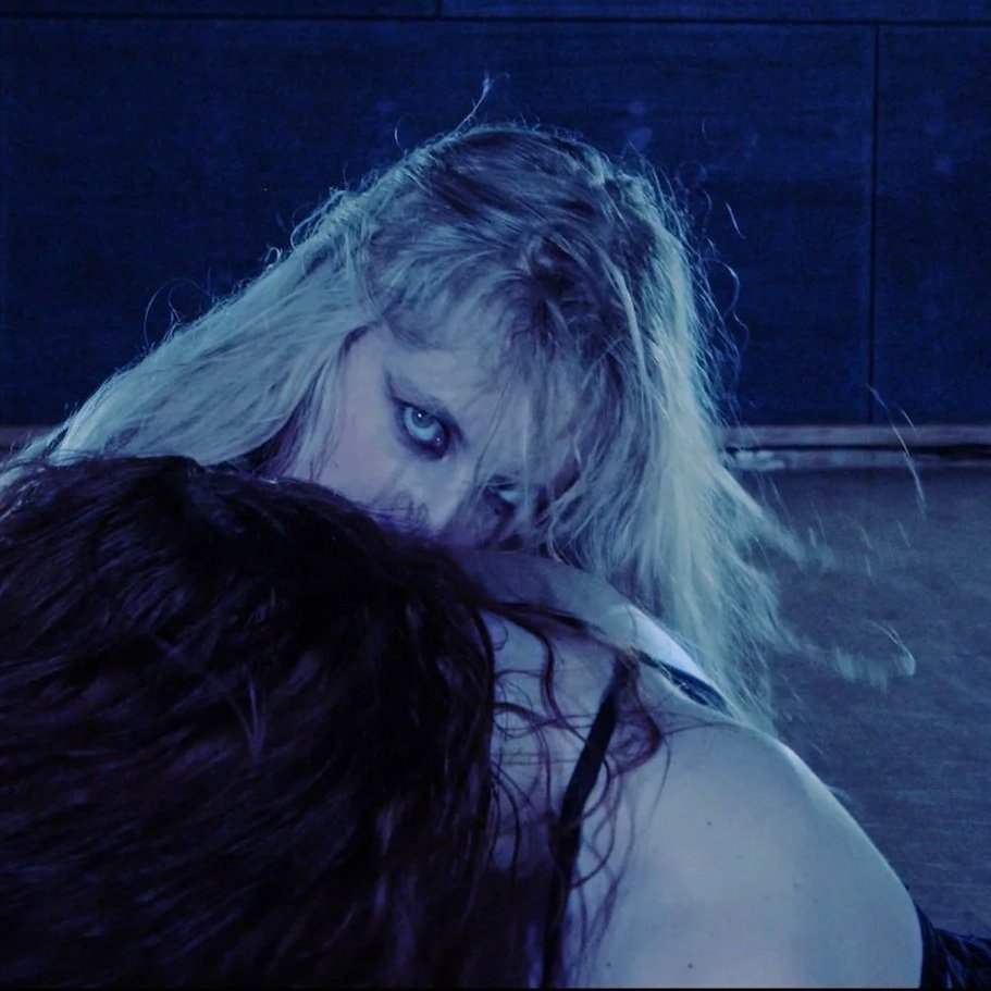 A woman with long, blonde hair and blue eyes looking intensely over her shoulder at the camera, with dark background and low lighting.