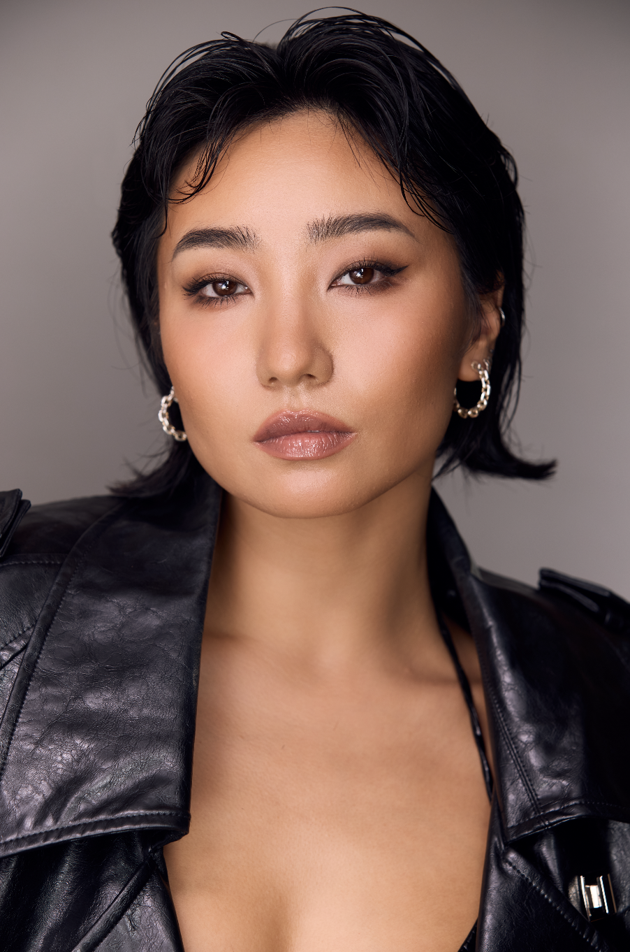 A woman with short black hair styled with wet look, wearing a black leather jacket, hoop earrings, and neutral makeup, looking directly at the camera.