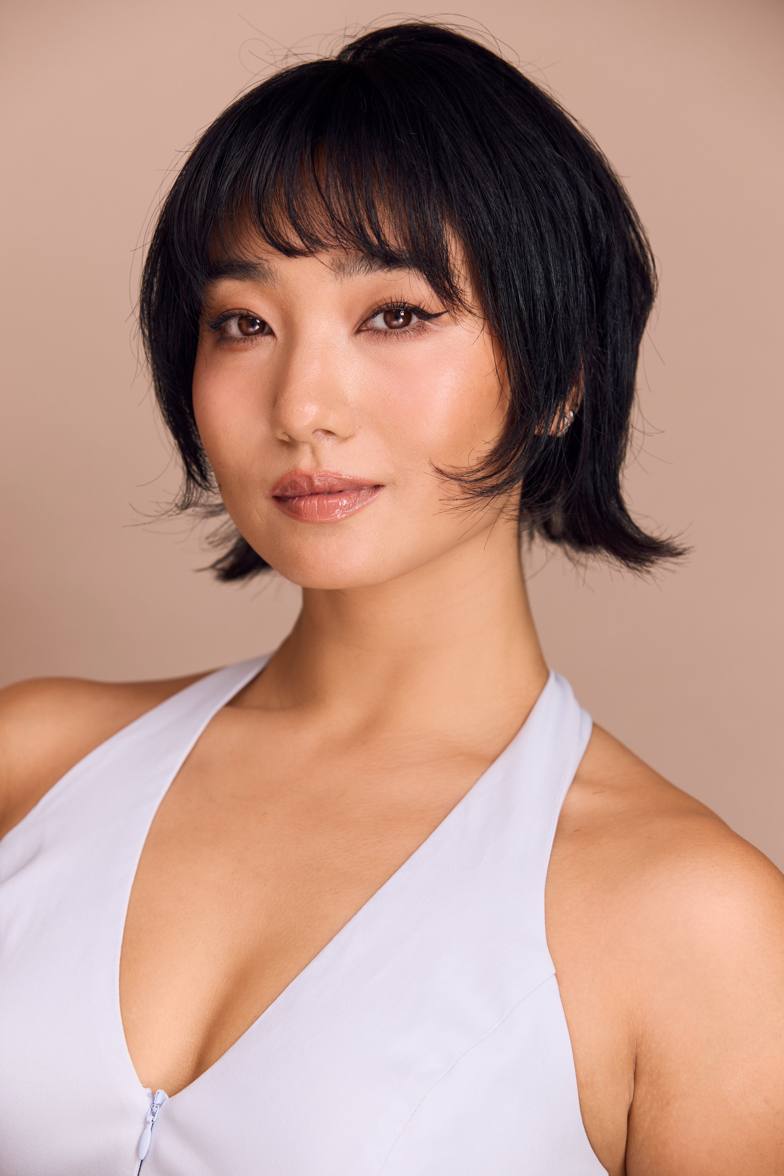 A close-up portrait of a woman with short black hair, wearing a white sleeveless top with a zipper, against a beige background.