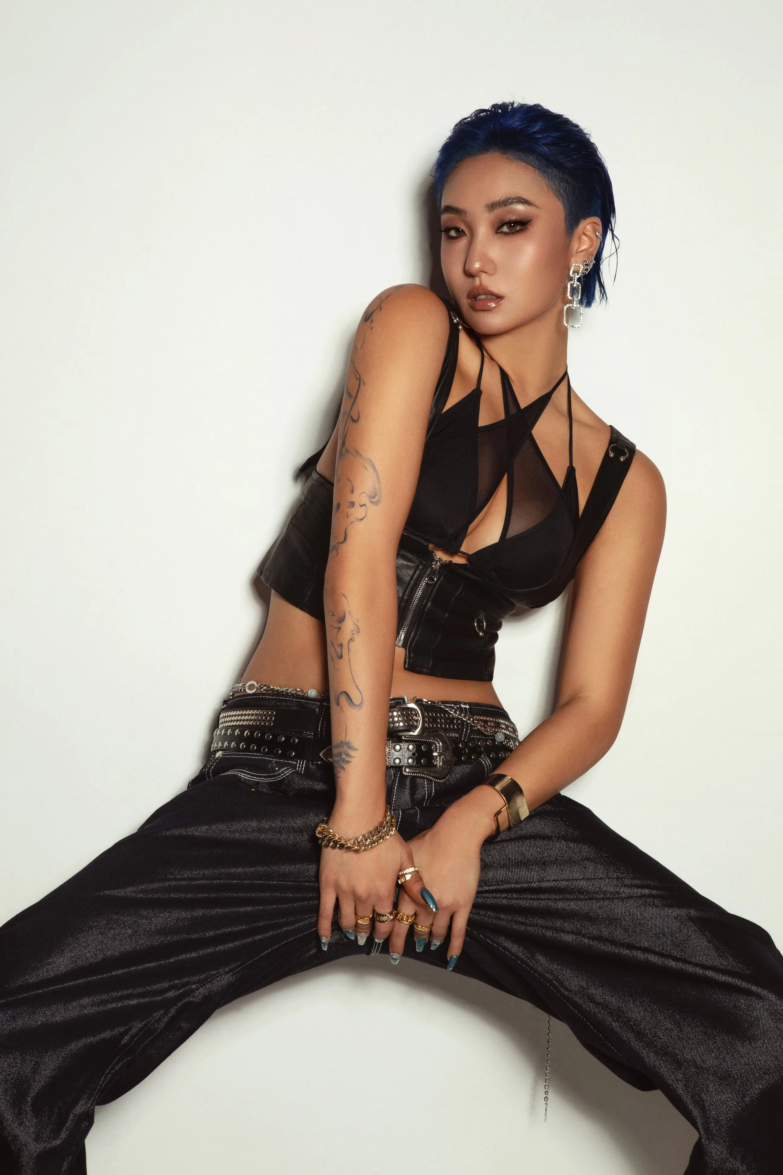 A woman with blue hair and a fierce expression wearing a black mesh top and baggy black pants, sitting with legs apart, against a plain white background, adorned with jewelry and tattoos.