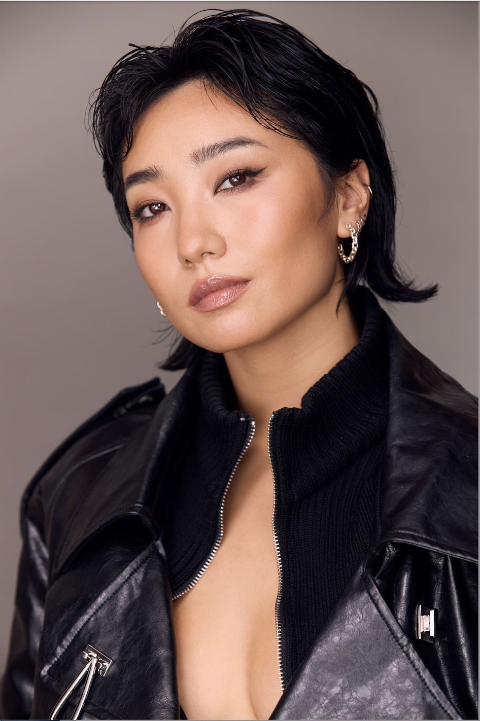 A woman with short black hair styled in a messy bob, wearing makeup with winged eyeliner and natural lipstick, dressed in a black leather jacket and black zip-up sweater, accessorized with multiple earrings, posing against a plain gray background.