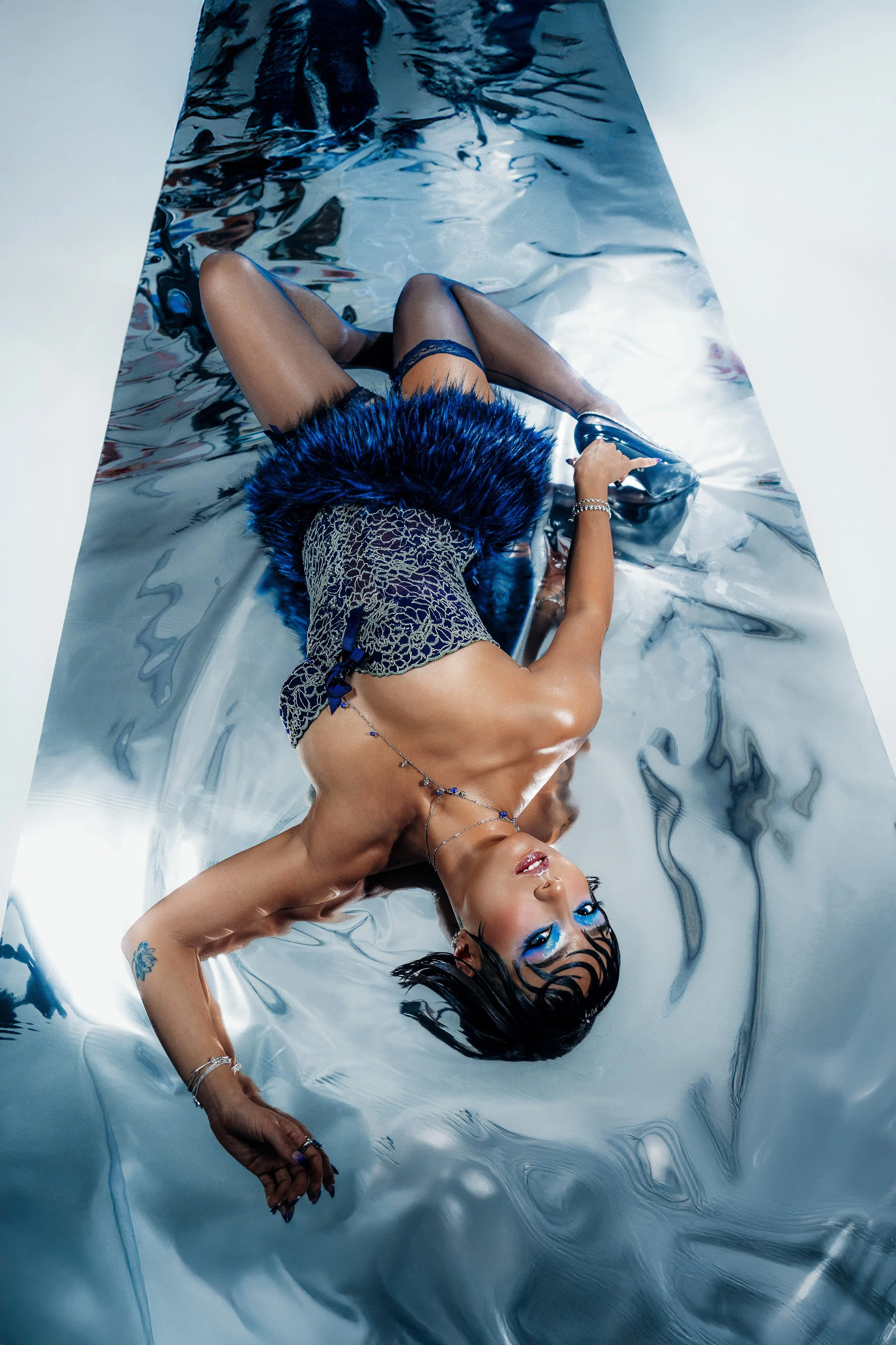 A woman lying on a reflective silver surface. She is wearing a leopard print top with blue accents, sheer black stockings, and high-heeled shoes. Her makeup is bold with blue eyeshadow and black eyeliner, and she has wet, black hair. She has tattoos and jewelry, including bracelets and a necklace.